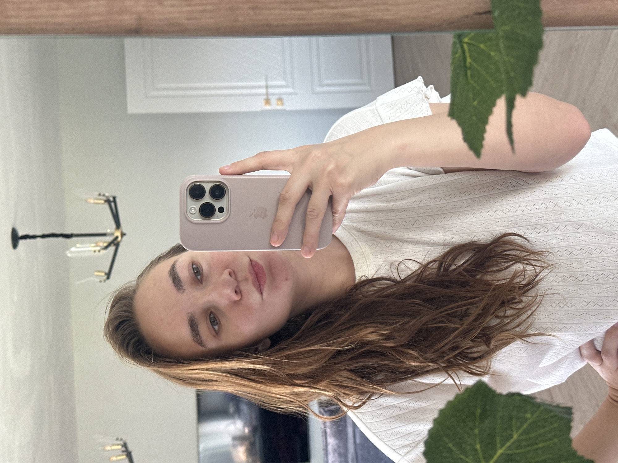 A person with light brown hair and fair skin is taking a selfie in a mirror. The person is wearing a white top. The reflection in the mirror reveals a chandelier, a door, and a television. A green leaf is visible in the foreground, partially obstructing the view. There is no information about Kharnavy, Russia, or its location in the image.