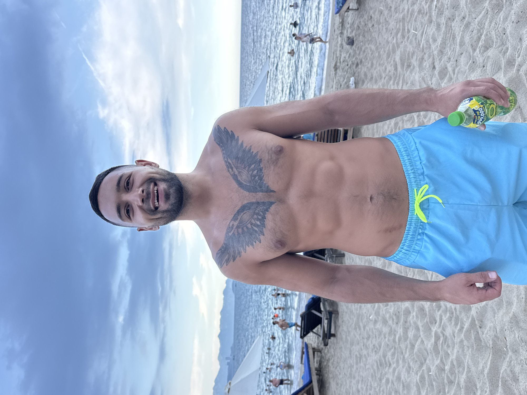A man stands on a sandy beach in Nha Trang Ward, Vietnam, holding a bottle of beverage. He is shirtless, wearing light blue swim trunks with a neon yellow drawstring. A large, dark tattoo of wings is visible on his chest. The man smiles at the camera. The background shows the ocean with people swimming and wading, lounge chairs, and umbrellas. In the distance, mountains and buildings are visible. The sky is overcast with clouds.