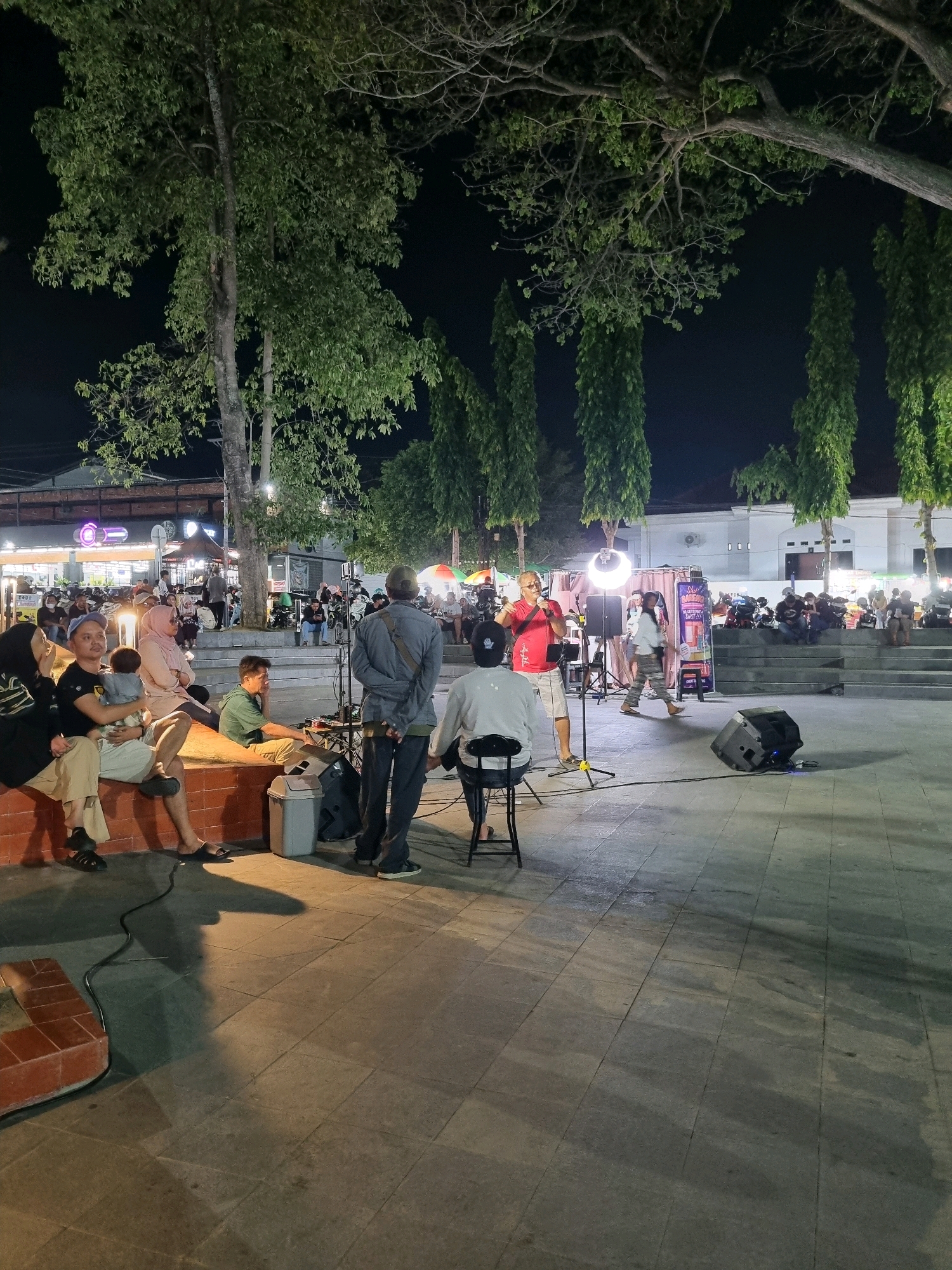 In Tanamodindi, Indonesia, at night, a group of people are gathered outdoors in what appears to be a public plaza or park. Several tall, slender trees with dense green foliage are visible, their leaves illuminated by artificial lighting. In the foreground, a man in a red shirt and shorts is holding a microphone and appears to be performing or speaking, standing in front of a circular light fixture and sound equipment, including speakers. Other individuals are seated on a raised, brick-like structure to the left, observing the performance. Further in the background, more people are seated, and there are stalls or shops with bright lights, suggesting a marketplace or event area. The ground is paved with large, square tiles. The overall atmosphere is that of a community gathering or outdoor entertainment event.