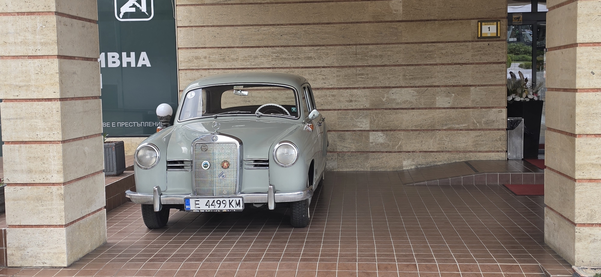 A light grey vintage Mercedes-Benz Ponton-series car (likely a W120 or W121 model from the 1950s) is parked under a covered entrance of a building in Sandanski, Bulgaria. The car is positioned centrally in the frame, facing forward. Its white steering wheel is visible through the windshield. Two circular badges adorn the grille below the Mercedes-Benz star: one depicting a mountain landscape and another a crest or emblem.
The car's Bulgarian license plate reads "BG E 4499 KM", with "РЕТРО ТИМ" (Retro Team) visible on the plate frame, suggesting it belongs to a classic car club.
The building's facade features textured light brown/beige stone cladding with darker horizontal lines, supported by two large, square pillars. The ground is covered with reddish-brown square tiles. To the left of the car, a glass door or window reflects greenery. On the right, another glass door leads into the building, with a numerical sign "1" above it. Inside, a decorative planter with flowers and a small waste bin are visible. Red mats are placed on the tiled floor near the entrance, which includes both a sloped ramp and steps.
Visible on the left glass facade, written in Cyrillic, are the words "ИВНА" and "ВЕ Е ПРЕСТЪПЛЕНИЕ", which are parts of a larger text that translates to "THE USE OF A COMMON WEAPON IS A CRIME". A stylized "A" or "A1" logo is also partially visible above "ИВНА".
The scene is set during the daytime under even lighting, with no strong shadows, suggesting an overcast day or shelter from direct sunlight. No people are visible, and no specific activity is taking place beyond the car being parked.