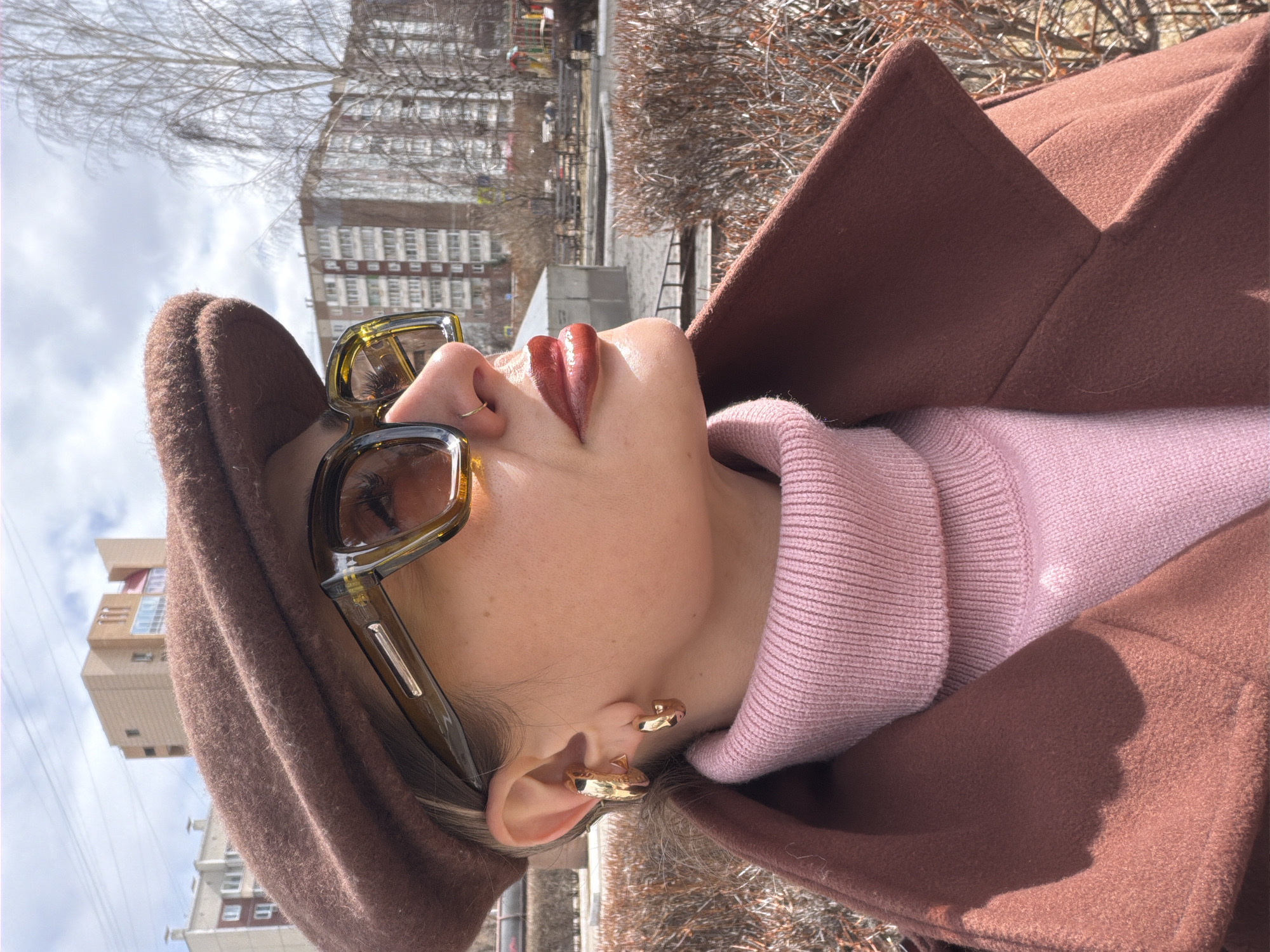 An upward-facing, close-up shot shows a person's profile from the nose up in an urban outdoor setting, likely Krasnoyarsk, Russia. The subject wears large olive-framed sunglasses, a small gold nose ring, and chunky gold ear cuffs on their visible right ear. Their lips are painted with a glossy red lipstick. They are dressed in a pink ribbed turtleneck sweater, over which a brown wool-like coat is worn, with a matching brown textured cap or hat partially visible on their head.
The person is looking upwards, suggesting they are observing the sky or surrounding architecture. The background features bare tree branches, indicating autumn, winter, or early spring. Several multi-story residential buildings with multiple windows are visible under a partly cloudy sky. The lighting suggests it is daytime, with shadows indicating direct sunlight. No discernible activity is taking place beyond posing or observation. There is no visible text in the image.