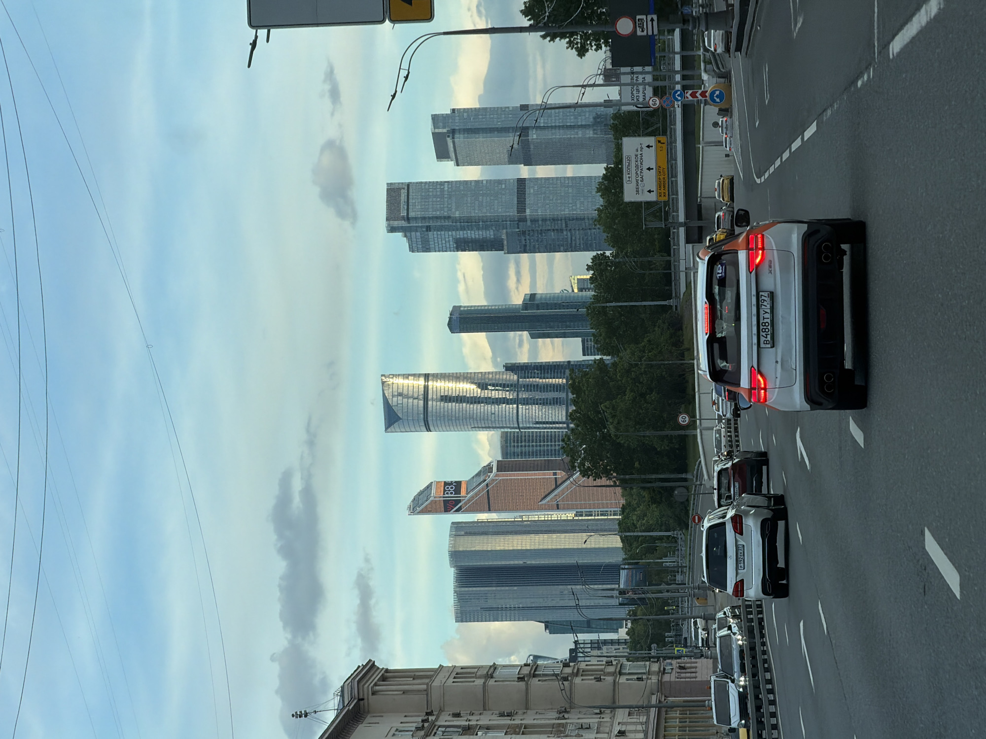 The image depicts a northbound traffic scene in Moscow, Russia. The Moscow International Business Center, also known as Moscow City, dominates the skyline in the distance, featuring several prominent skyscrapers. A multi-lane road stretches into the distance, filled with cars. Traffic signs are visible alongside the road, indicating directions to the 3rd Ring Road, Zvenigorodskoe Highway, and other destinations, with speed limits also indicated. The license plate of a car in front is visible, showing "B488TY 797", while another nearby car's plate reads "P192 EK 77". The sky is partly cloudy. A building with rooftop features and windows is seen in the foreground on the left side of the road.