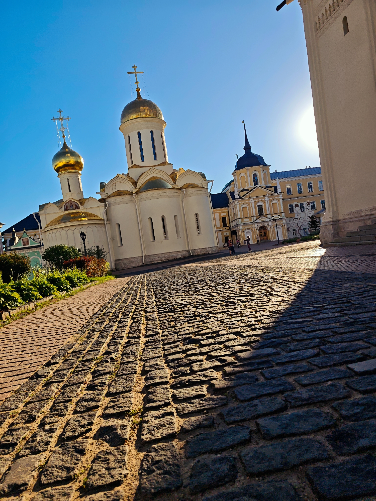 The original image displays a cobblestone path leading towards a religious complex under a clear blue sky. The sun is shining brightly, casting long shadows on the path. On the left side of the path, there are lush green bushes and some red foliage. The religious complex features several buildings with white walls and golden domes topped with crosses, characteristic of Orthodox Christian architecture. One prominent building has two golden domes and an arched facade. To the right, a taller, light-colored structure with decorative elements is partially visible, along with a bright yellow building adorned with a dark steeple. A few people are visible in the distance, walking in front of the yellow building. The scene appears to be a sunny day in an urban or historical setting, possibly a monastery or church compound.