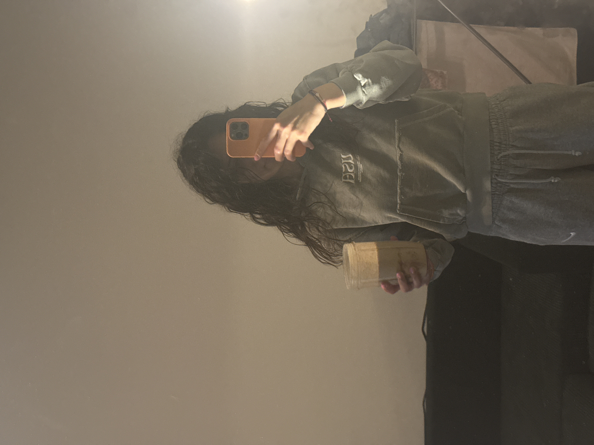 A young person with long, dark, wet hair is holding an orange smartphone in their left hand, taking a selfie in a mirror. The person is wearing a gray hoodie and matching sweatpants. The reflection in the mirror shows a glimpse of the room, including a canvas on a stand and dark curtains or a dark wall. The person is holding a clear jar filled with a light-brown or tan substance in their right hand. There is no indication of the location being Sofia, Bulgaria.
