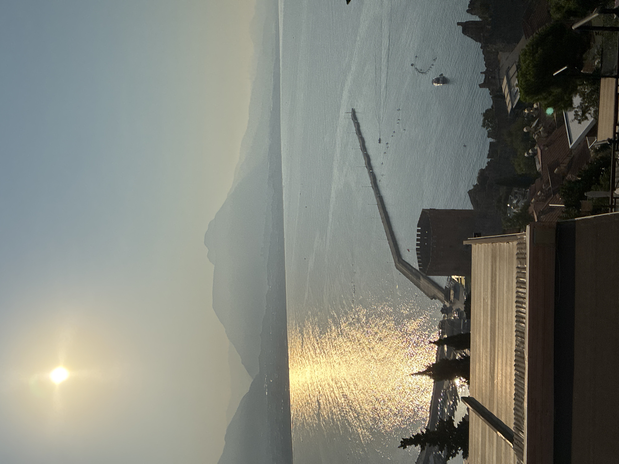 This is an outdoor, high-angle, medium shot photograph of the sea and coastline of Alanya, Turkey, taken during the day. The sun, positioned in the upper left quadrant of the image, casts a strong glare on the water, creating a shimmering effect. A long, narrow pier extends out into the sea from the right side of the frame. Several boats, including a white motorboat, are visible on the water, along with what appear to be paddleboarders. A red, circular tower, possibly a defensive structure, is situated on the coast near the pier. In the foreground, the rooftop of a building is visible, constructed with light-colored, ribbed panels. The background features hazy mountains, suggesting a slightly overcast or dusty atmosphere. The overall scene depicts a coastal landscape with elements of human activity and historical architecture.
