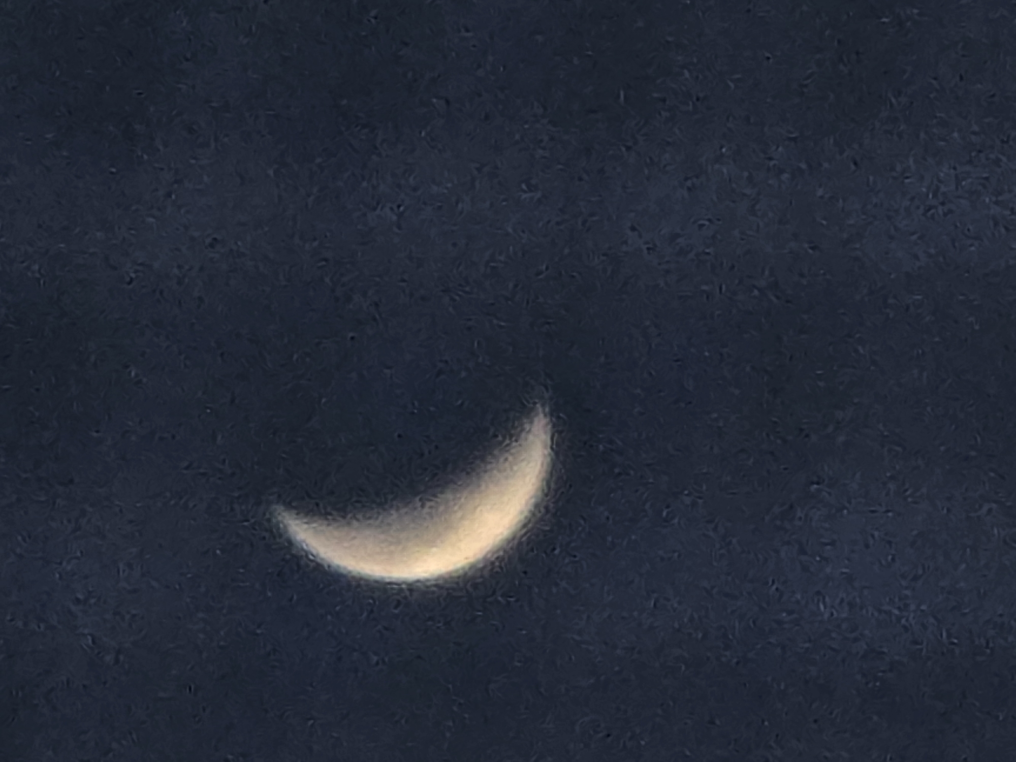 A crescent moon is visible in a dark blue sky. The moon is illuminated from the right, with its left side appearing darker. The sky has a textured appearance, with subtle variations in darkness that suggest a night sky. The image appears to be a photograph taken at night. There are no people, objects, or actions depicted other than the moon and sky. The location in Ермолаево, Russia is not discernible from the image.