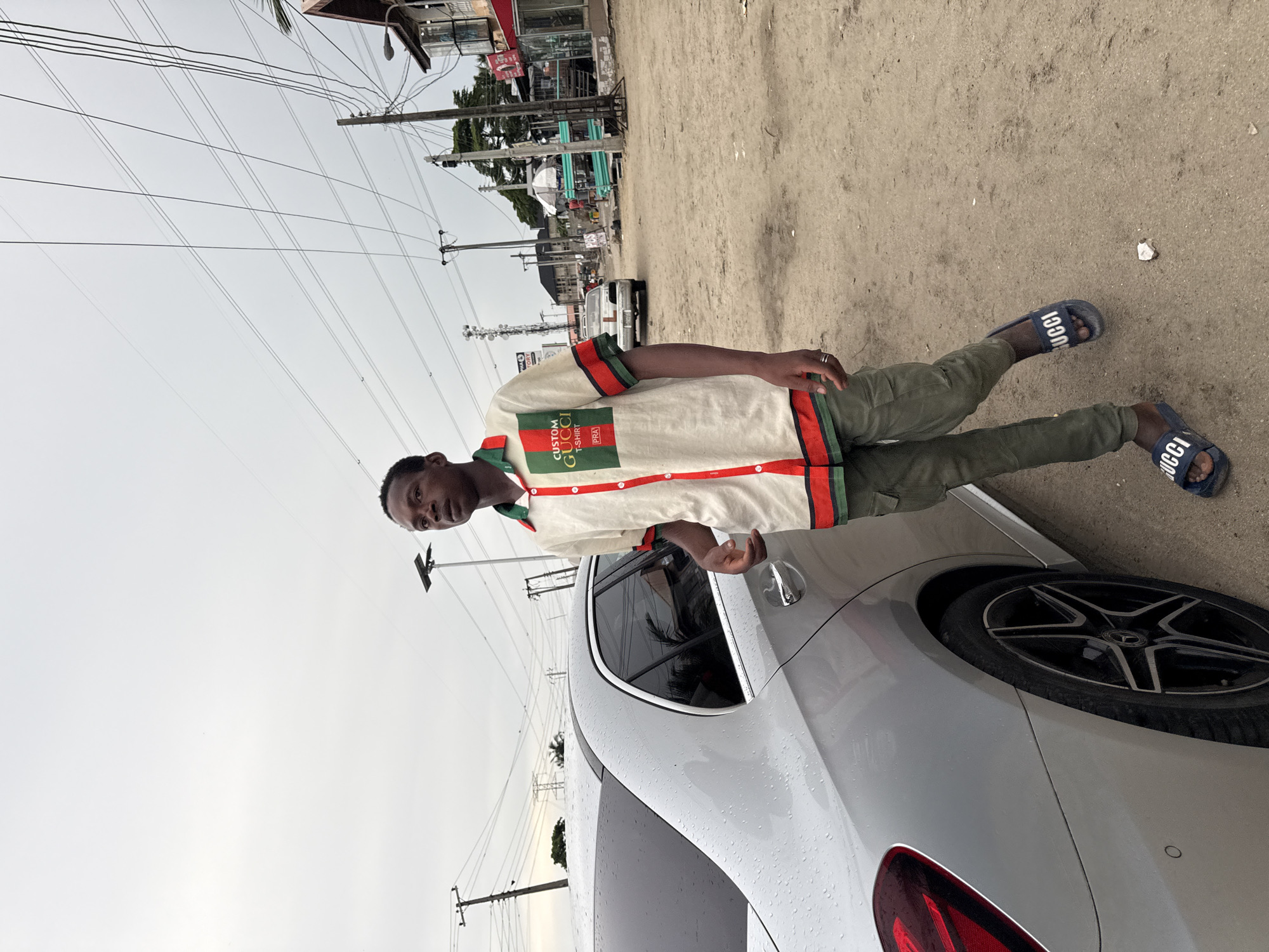 A young man stands near the rear of a silver vehicle on a dirt or sandy shoulder of a road in Ado, Nigeria. He wears a light-colored, button-down shirt with red and green stripes, olive green pants, and blue "Gucci" slides. The vehicle appears to be a modern sedan with alloy wheels and a partially visible tail light. Behind the man and vehicle is a street with shops and utility poles, typical of an urban or semi-urban environment. The sky is overcast.