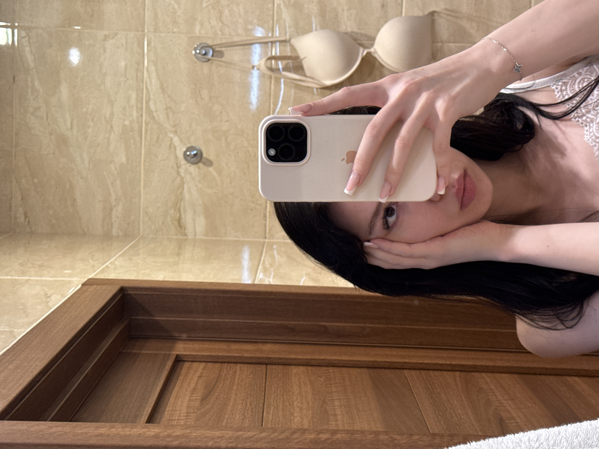 In a bathroom, a young woman with long, dark hair takes a selfie in a mirror. She is holding a light-colored smartphone in her right hand, partially obscuring her face. Her left hand is placed on her cheek. She is wearing a white lace camisole. Behind her, on the tiled wall, a beige bra is hanging from hooks. A wooden door is visible on the left side of the frame. The scene appears to be in an apartment bathroom, possibly in Khimki, Russia, given the context.