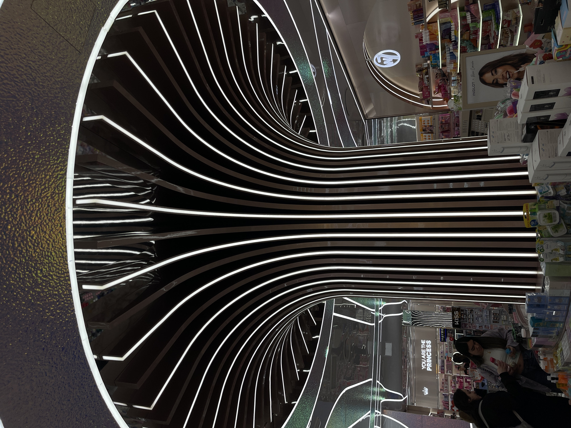 The image depicts the interior of a modern beauty and cosmetics retail store, likely a Primor store in Valencia, Spain, characterized by its distinctive architectural design and visible brand signage.
At the center of the frame, a large, inverted, tree-like structure serves as a central design element. This structure features numerous dark, reflective panels, each illuminated along its edge by a bright white LED strip. These strips curve upwards and outwards, forming a multi-ribbed, glowing canopy that extends towards the edges of the image. The ceiling directly above this structure has a textured, reflective surface with a golden-brown, dimpled pattern.
The store's retail environment is visible around this central feature. On the left, two adult females are interacting with products displayed on a counter. The woman on the right, wearing a light top, a striped open shirt, and jeans, holds a pink bottle in her right hand and a blue bottle in her left, appearing to examine them. The woman to her left is partially obscured, wearing dark attire and looking down at the counter. In the background behind them, a white sign reads "YOU ARE THE PRINCESS" accompanied by a crown emblem, above shelves stocked with various products. Other visible brand names, partially obscured, include "MAYBE," "essie," and "KISS."
On the right side of the image, another display unit features a circular "W7" logo above multiple shelves densely packed with colorful beauty products. Below this, a promotional poster for "INGLOT x Rocío Camacho" displays a smiling woman. Various boxed products, including what appear to be skincare, cosmetics, and household items (some in green packaging), are visible on display tables and shelves in the foreground and background. The overall lighting is bright and even, emanating primarily from the architectural LED strips and ambient store fixtures.