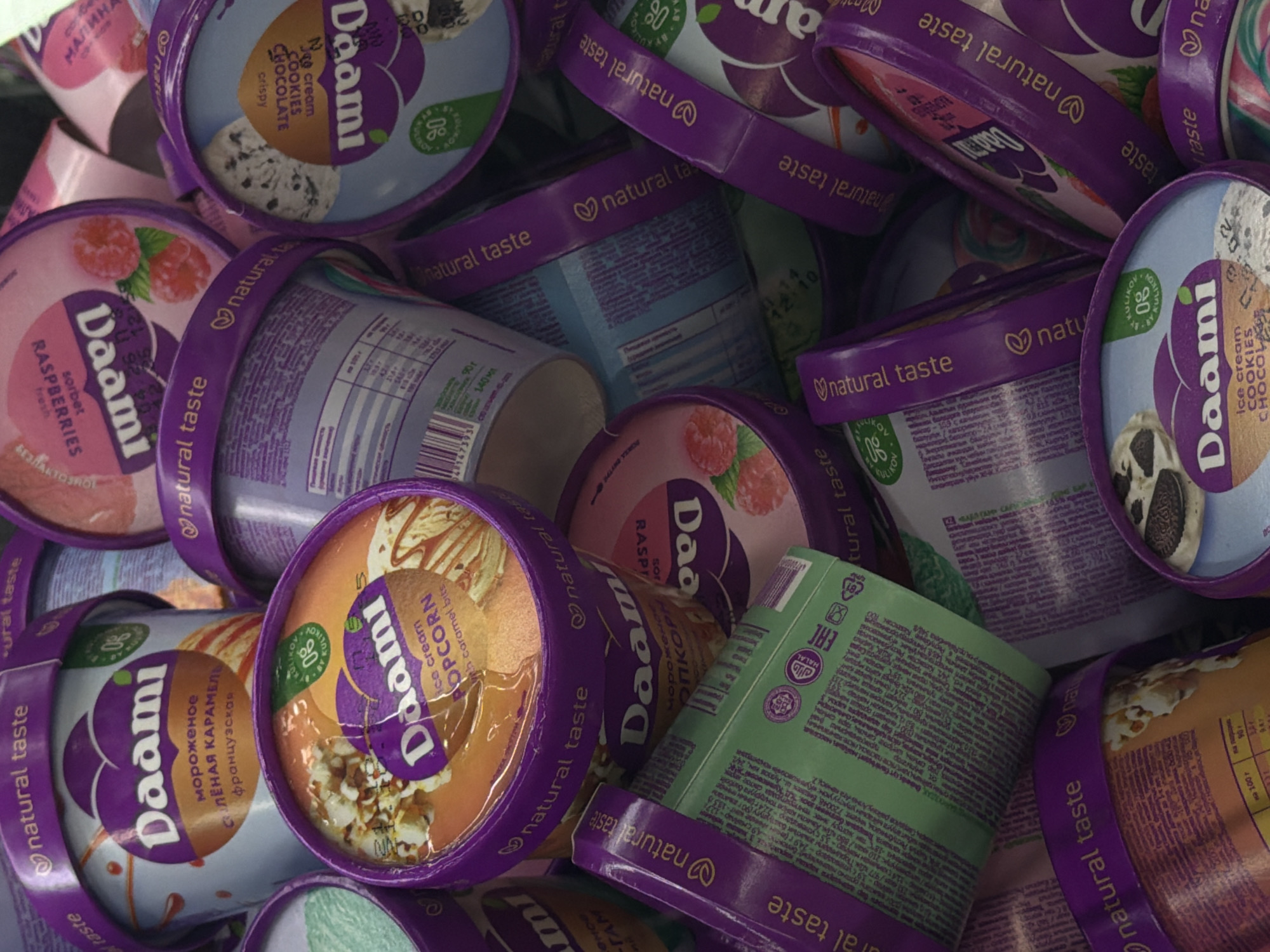 The image displays a dense, haphazard pile of numerous "Daami" brand ice cream containers, captured from a high-angle, close-up perspective. The containers are predominantly cylindrical, featuring purple lids with a "natural taste" label and a heart-shaped logo. The container bodies vary in color, including light gray, orange, pink, light blue, and green.
Visible flavors include "salted French caramel," "ice cream popcorn with caramel bites," "raspberries sorbet," and "ice cream cookies chocolate" with chocolate chips. Many lids feature a "0% Kulikov" emblem. A green-colored container clearly displays a Halal certification logo and an EAC (Eurasian Conformity) mark. Several containers reveal barcodes and nutritional information tables on their sides.
The containers are stacked randomly, with some upright, others tilted, and some inverted, creating a cluttered arrangement that obscures the immediate background. The scene is brightly lit, causing reflections on the container lids. The specific location within the city of Unknown, Unknown, is not discernible. No people or actions are depicted.