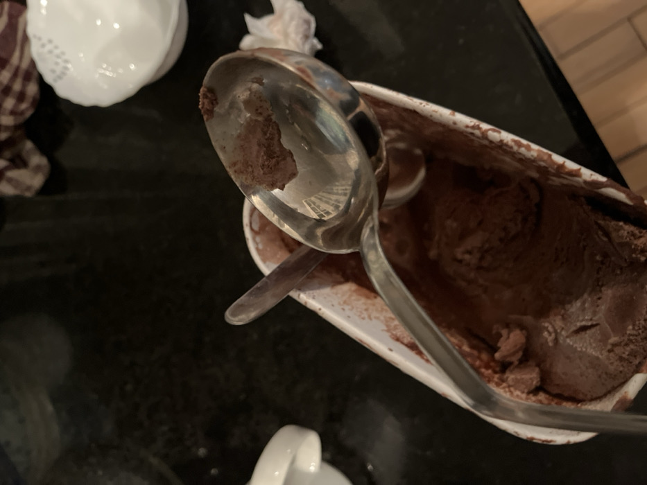 The scene depicts a partially consumed rectangular white cardboard container of chocolate ice cream situated on a dark, reflective countertop in a kitchen in Joliet, United States of America. Inside the container, a large, reflective silver metal ice cream scoop rests with its bowl facing upwards, containing a small amount of chocolate ice cream residue. A smaller, dark-colored spoon is also present within the container, partially submerged in the ice cream. The chocolate ice cream shows signs of having been scooped, with an uneven, slightly softened surface. In the upper right background, a white ceramic bowl with a textured rim is visible, along with a brown and white checkered cloth. A partial view of another white ceramic object is present in the upper left background. No individuals are visible.