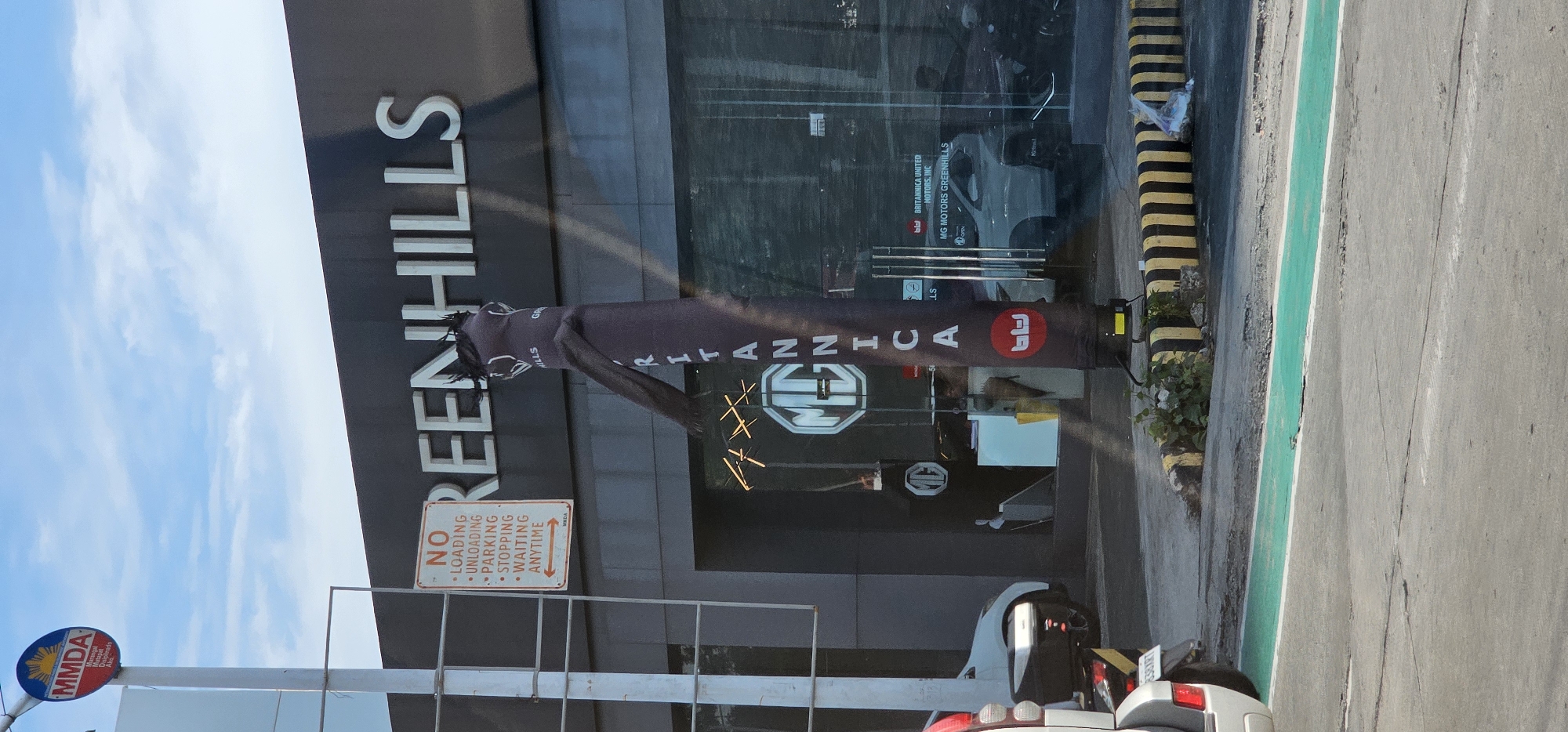 The image captures an outdoor scene in front of a car dealership, specifically MG Motors Greenhills in San Juan, Philippines. The prominent signage above the building reads "GREENHILLS" in large, white, uppercase letters against a dark background. In the foreground, a dark-colored inflatable tube man, a common advertising tool, stands tall, its fabric body twisted by the wind. The tube man has "BRITANNICA" written vertically in white letters.
To the left of the inflatable man, a white pole supports two signs. The upper sign is a circular MMDA (Metropolitan Manila Development Authority) logo with a sun emblem, and text below it. The lower sign is an orange and white rectangular sign with "NO LOADING, UNLOADING, PARKING, STOPPING, WAITING ANYTIME" printed in black text. An orange arrow points to the right.
The dealership's entrance features large glass doors with prominent MG logos visible inside. Through the glass, several MG cars are displayed. To the far left, the rear end of a white vehicle and a part of a silver SUV are visible, suggesting traffic or parking nearby. The ground is paved, with a strip of bright green paint marking a lane or path. Yellow and black striped bollards line the edge of the sidewalk, and some vegetation grows around them. The sky above is blue with scattered white clouds. The overall impression is that of a commercial street view, likely during daylight hours.