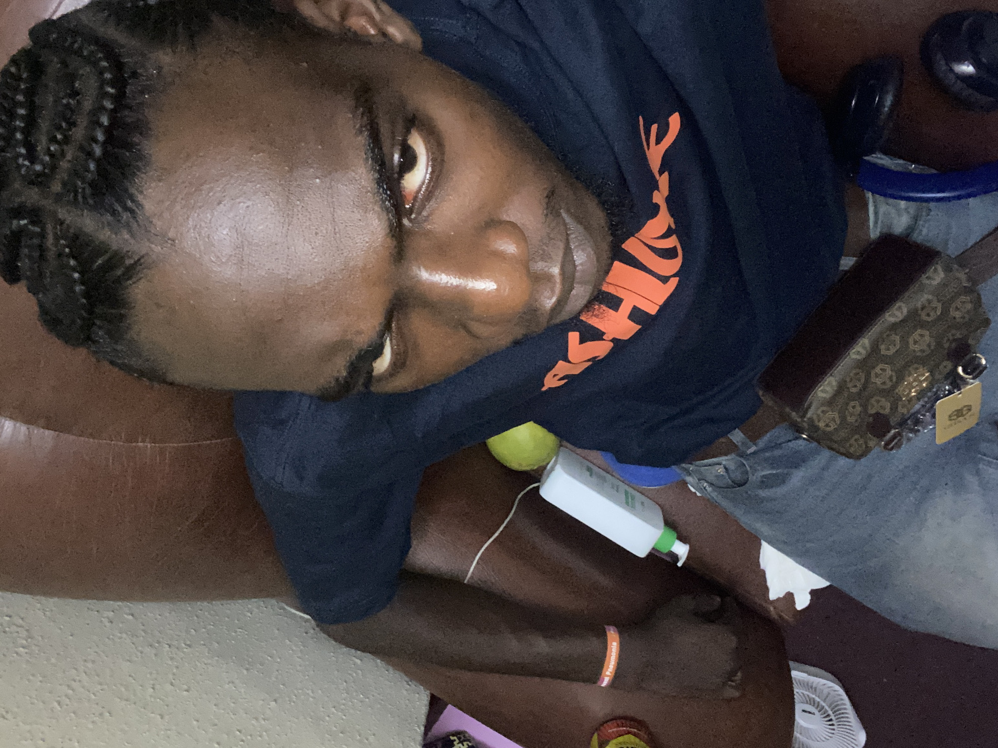 An adult male with dark complexion and tightly braided cornrows is depicted from a high-angle perspective, gazing directly into the camera. He is seated or reclining in a brown, worn, leather-like armchair within an indoor setting in Lagos, Nigeria. His forehead is shiny, and both eyes show a reddish tint in the sclera. He wears a navy blue t-shirt with a partially visible orange graphic and light blue denim jeans. An orange "Treat Pneumonia" wristband is on his left wrist. A patterned brown fanny pack or small crossbody bag with a gold-colored tag rests across his lap. On the chair to his left, a red and yellow container and a small white electric fan are visible. To his right, on the chair, are a white pump-dispenser bottle, a yellow-green spherical fruit, and a thin white cord. A crumpled white tissue lies near his left thigh. A blue curved object and a dark circular item are visible on the floor near his right leg. The background reveals an off-white, textured wall behind his left shoulder.