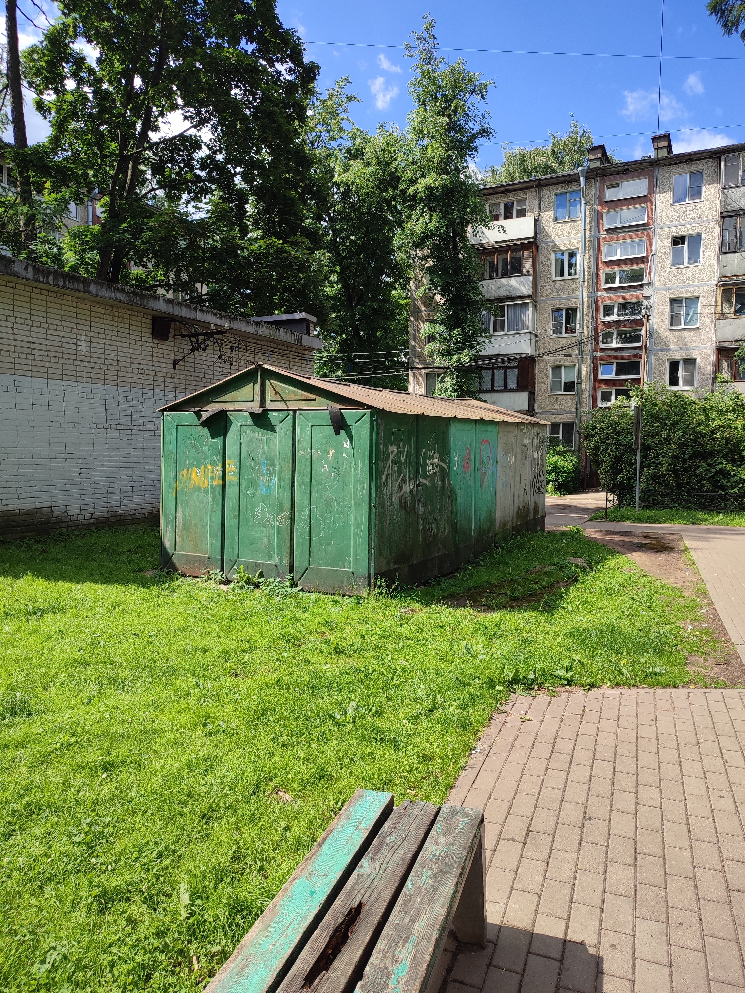 The image depicts a green, graffiti-covered shed in an urban setting. The shed has a metal roof and appears to be made of corrugated metal panels. It is situated on a patch of green grass with weeds growing around its base. To the left of the shed is a white brick wall. Behind the shed and to the right, a multi-story apartment building with a beige facade and multiple windows is visible, partially obscured by trees. A paved walkway with brick pavers runs along the right side of the image, and a wooden bench with peeling green paint is in the foreground. The sky is blue with scattered white clouds. The scene appears to be from Vsevolozhsk, Russia, given the context.