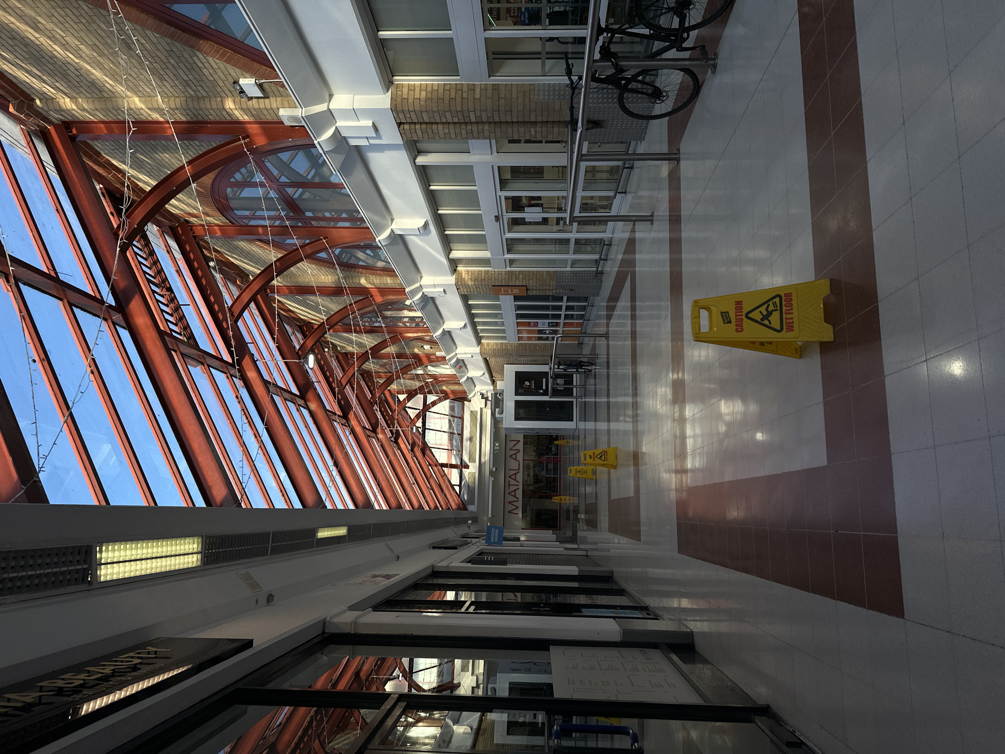 The image shows an indoor walkway in a shopping center or mall. The walkway has a high, arched glass roof supported by red metal beams. The walls are lined with storefronts with glass windows and doors. A "CAUTION WET FLOOR" sign is prominently placed in the center of the walkway. A bicycle is parked nearby. The floor is made of light-colored tiles with reddish-brown accents. A store called "MATALAN" is visible in the background. It is not possible to determine the exact location in the city of London, United Kingdom from the image.