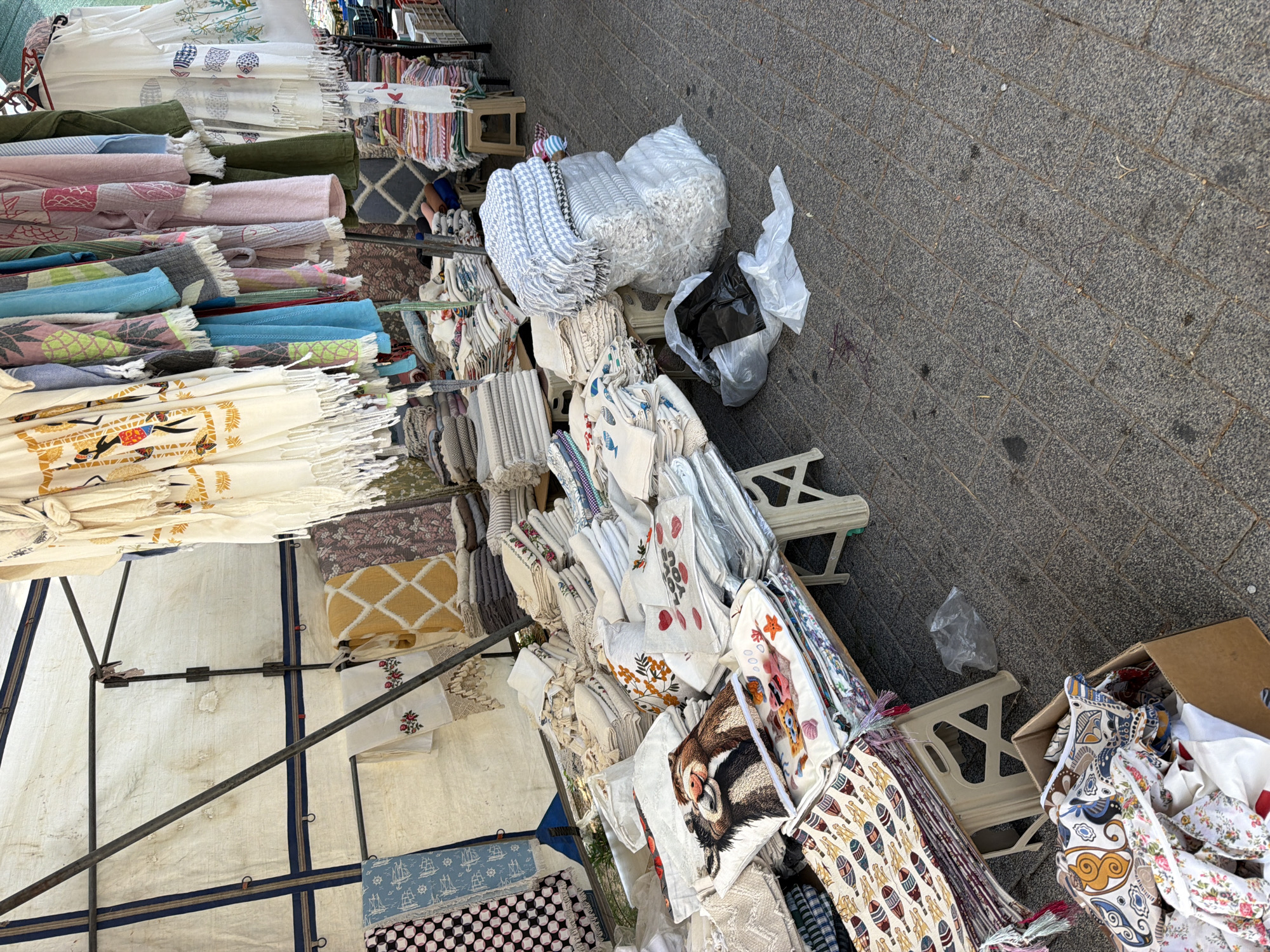 A street market stall in Bodrum, Turkey, displays a variety of textiles, including blankets, throws, and decorative pillows. The stall features a white canvas awning supported by metal poles. Various stacks of folded fabrics are arranged on tables and on the ground. Some items are patterned with traditional Turkish motifs, while others feature cartoon characters or floral designs. One pillow prominently displays an image of two raccoons. In the foreground, a cardboard box overflows with more textiles. The ground is paved with gray cobblestones. No people are visible in the frame.