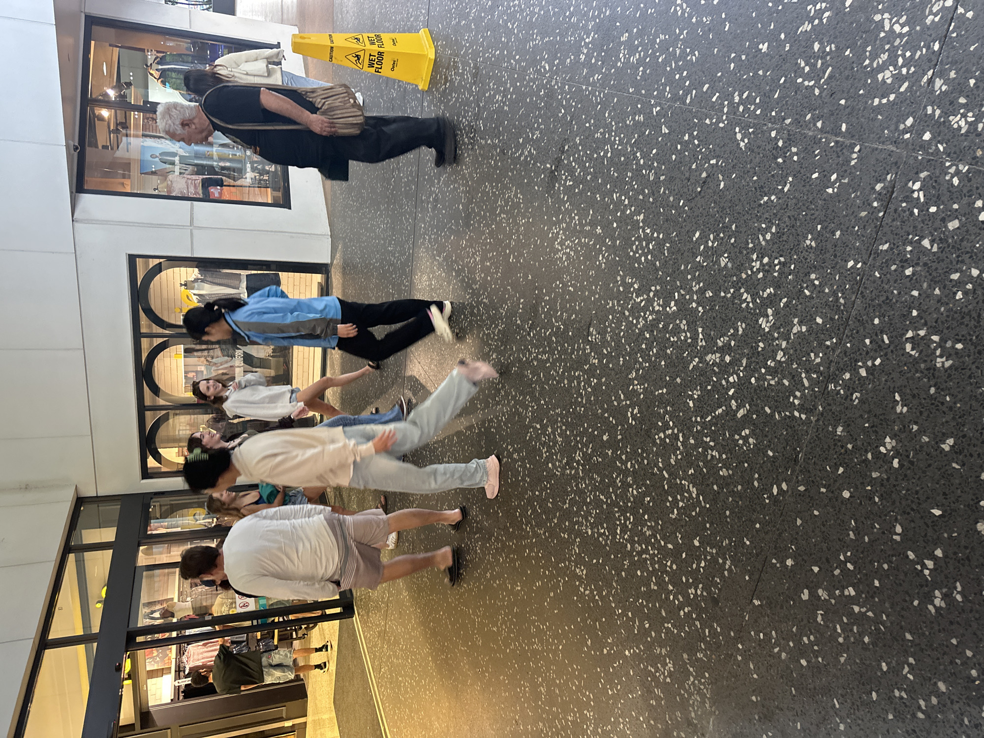 The image shows a group of people walking on a speckled dark grey tiled floor inside what appears to be a shopping center or mall. The floor has a pattern of white flecks. To the left, there is a shop entrance with glass doors and displays of clothing. Further to the right, a yellow "Wet Floor" caution cone is visible. The lighting suggests an indoor environment. The scene depicts everyday activity with individuals in casual attire.