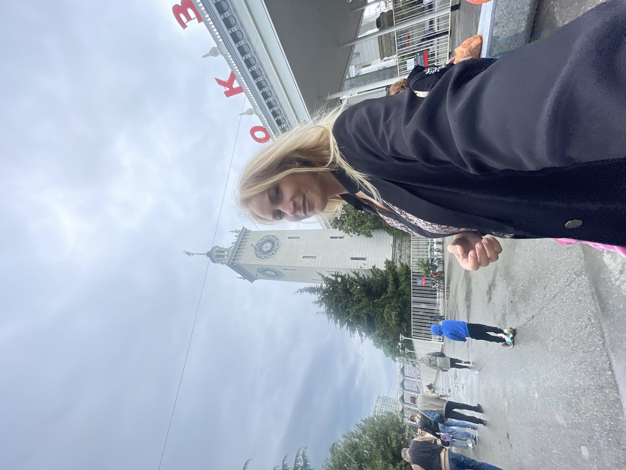 A medium shot captures a blonde woman, possibly taking a selfie, on a cloudy day in front of the Sochi Train Station clock tower in Sochi, Russia. The woman is wearing a black coat. In the background, people are walking on the wet pavement; one person is on a skateboard. The sky is overcast. Red letters are partially visible above the woman, likely part of a sign. The Sochi Train Station clock tower is visible behind the woman. A green evergreen tree is near the clock tower. Metal barriers are visible as well.