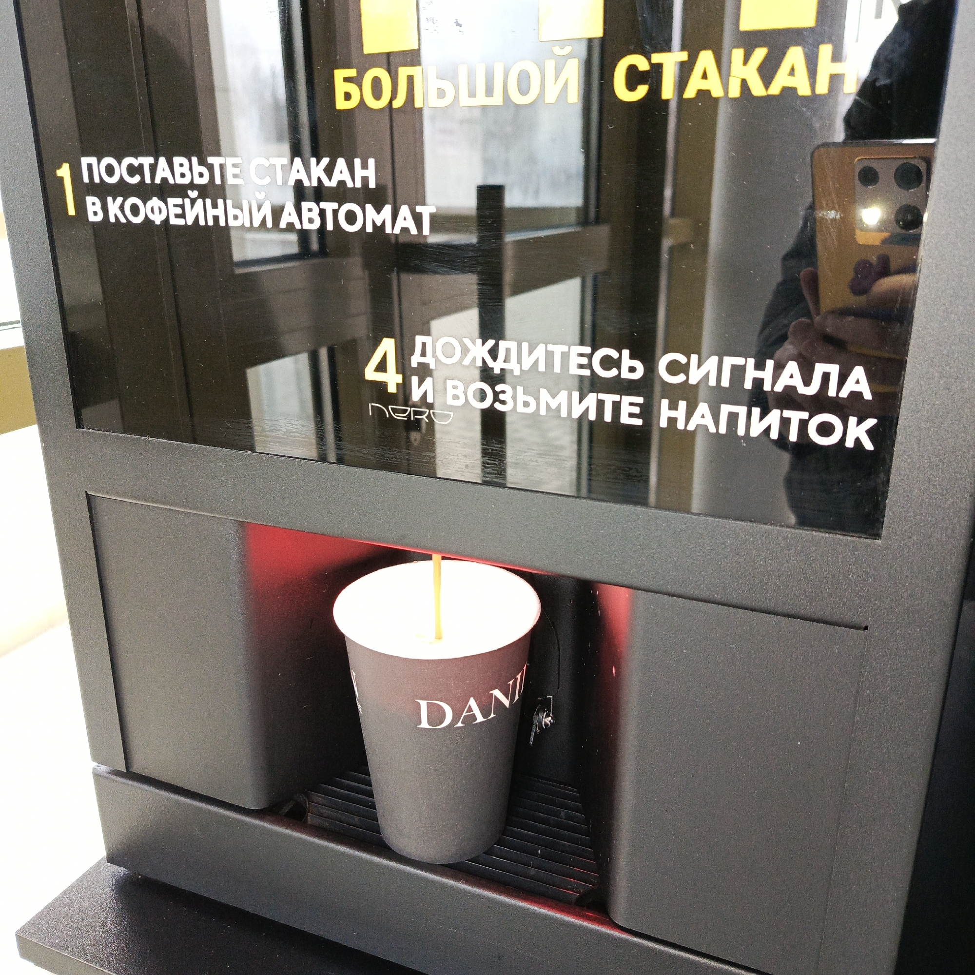 The image displays a close-up of a self-service beverage machine, likely a coffee dispenser, operating in Chaikovsky, Russia. The machine is black, featuring a glossy upper panel that serves as a display and a matte lower section where the drink is dispensed. A red light illuminates the internal area where a dark-colored disposable cup, branded "DANI" in white letters, is receiving a stream of light-colored liquid, presumably coffee or a similar hot beverage.
The glossy display panel contains instructions in Russian. Prominently at the top, yellow text reads "БОЛЬШОЙ СТАКАН" (LARGE CUP). Numbered white instructions guide the user: "1 ПОСТАВЬТЕ СТАКАН В КОФЕЙНЫЙ АВТОМАТ" (1 PLACE THE CUP IN THE COFFEE MACHINE) and "4 ДОЖДИТЕСЬ СИГНАЛА И ВОЗЬМИТЕ НАПИТОК" (4 WAIT FOR THE SIGNAL AND TAKE THE DRINK). The word "NERO" is also faintly visible in small letters near the fourth instruction.
Reflected in the machine's glossy surface is a person holding a smartphone, likely the photographer. The phone has a yellow or gold case, a visible multi-lens camera system, and a small heart-shaped sticker, indicating personal use. The setting appears to be an indoor public space, possibly an office, lobby, or a self-service station, given the functionality of the machine.