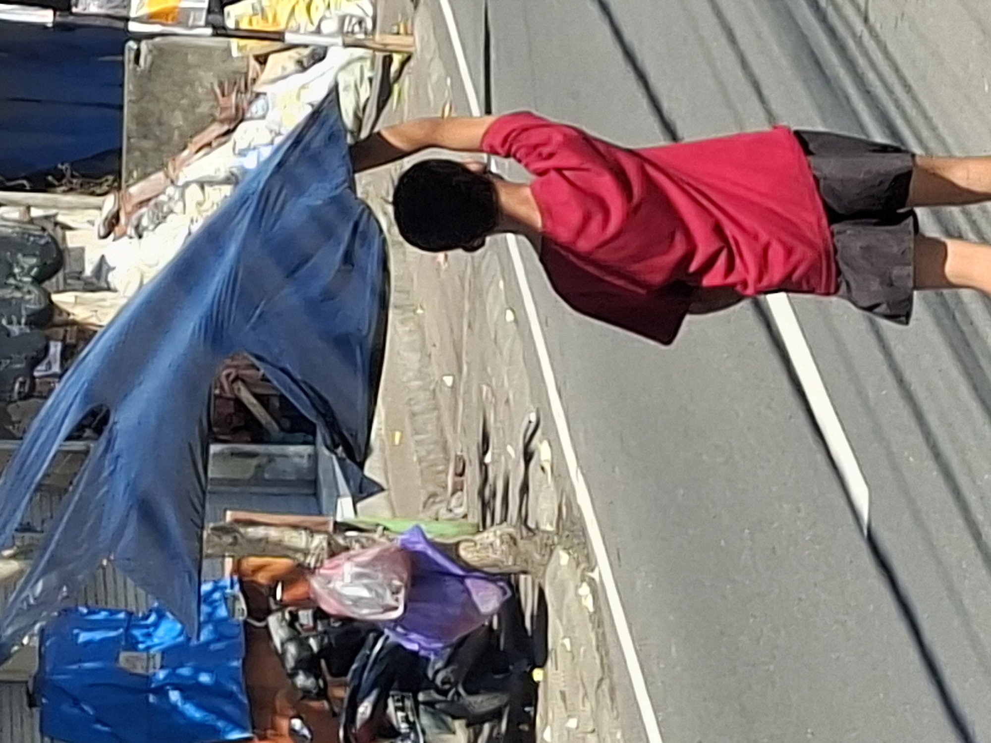 In Blahbatuh, Indonesia, a young person wearing a red shirt and black shorts walks on a street, holding a large, dark blue tarp above their head as a makeshift umbrella. The tarp is distorted by the wind, creating an abstract shape. Behind the person, a motorcycle is parked near a tree with bags, including a purple one and a clear plastic bag containing items, hanging from it. The environment suggests a street scene with some buildings and structures in the background, characterized by a somewhat cluttered and outdoor setting. The road has white markings, indicating it is a paved surface. The lighting suggests it is daytime.