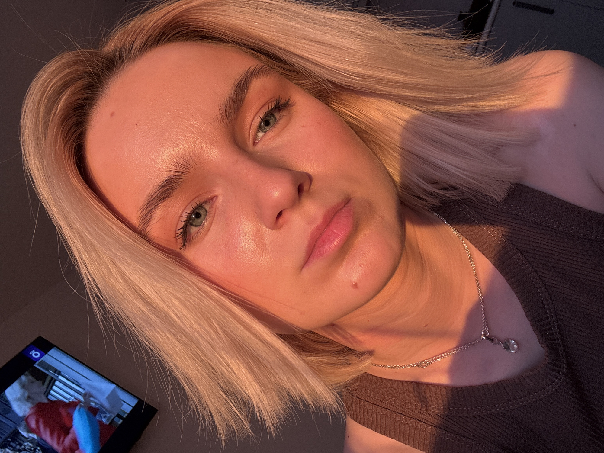 A close-up, slightly angled, indoor photograph shows a fair-skinned woman with short, blonde hair and green eyes. She is wearing a brown, ribbed tank top and a delicate silver necklace with a pendant. She appears to be illuminated by warm, golden sunlight. To the left of the frame, a small television screen is visible, showing what appears to be a program with a person in a red jacket. The overall setting and lack of specific landmarks make it impossible to ascertain the location within the city of Vsevolozhsk, Russia.