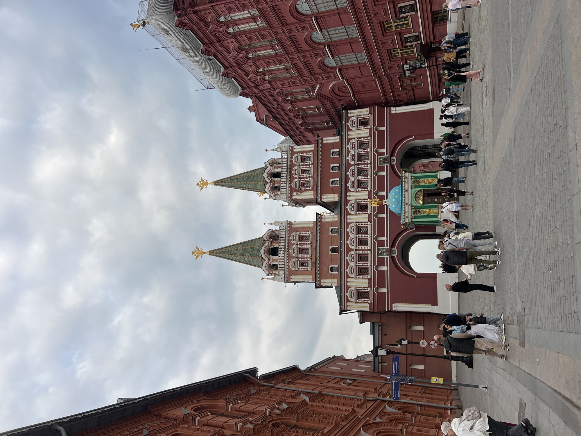 The image shows a street scene in Moscow, Russia, featuring the Resurrection Gate, a red brick structure with two towers topped with green spires and golden double-headed eagles. The towers flank an archway filled with people. Two red brick buildings frame the gate, one above and one below. The sky is overcast. People are walking and standing around.