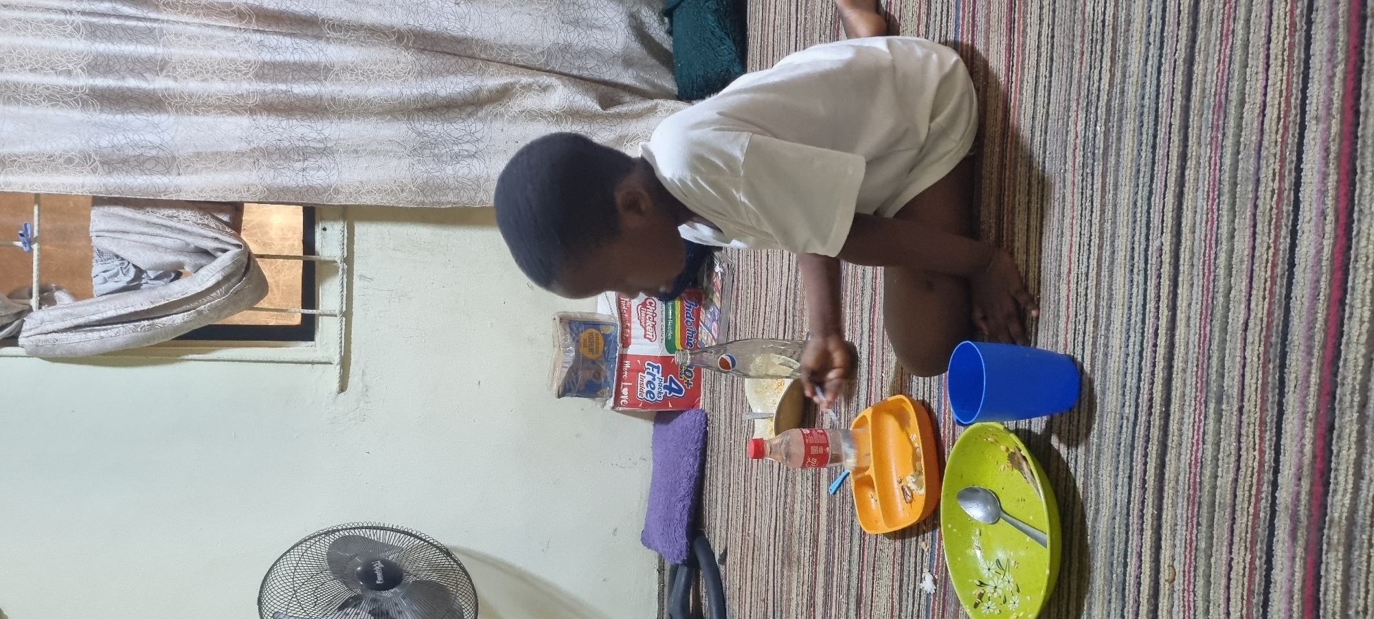 In Lagos, Nigeria, a young boy with short, dark hair sits cross-legged on a striped carpet, facing away from the camera and to the right. He is wearing a white t-shirt and appears to be eating. In front of him, arranged on the carpet, are several items. A green oval plate with a floral pattern contains leftover food and a spoon. Next to it is a blue plastic cup. Slightly further away, an orange compartmentalized plate also holds a small amount of food. A clear plastic bottle with a red cap, likely containing a beverage, stands upright. Behind the plates, a glass Pepsi bottle is visible, along with a few stacked food boxes. To the left of the boy, a black oscillating fan is positioned against a cream-colored wall. A purple rectangular cushion lies on the floor near the fan. In the background, a window is covered by a patterned curtain, and some light emanates from behind it. The overall scene depicts a simple mealtime in an indoor setting.