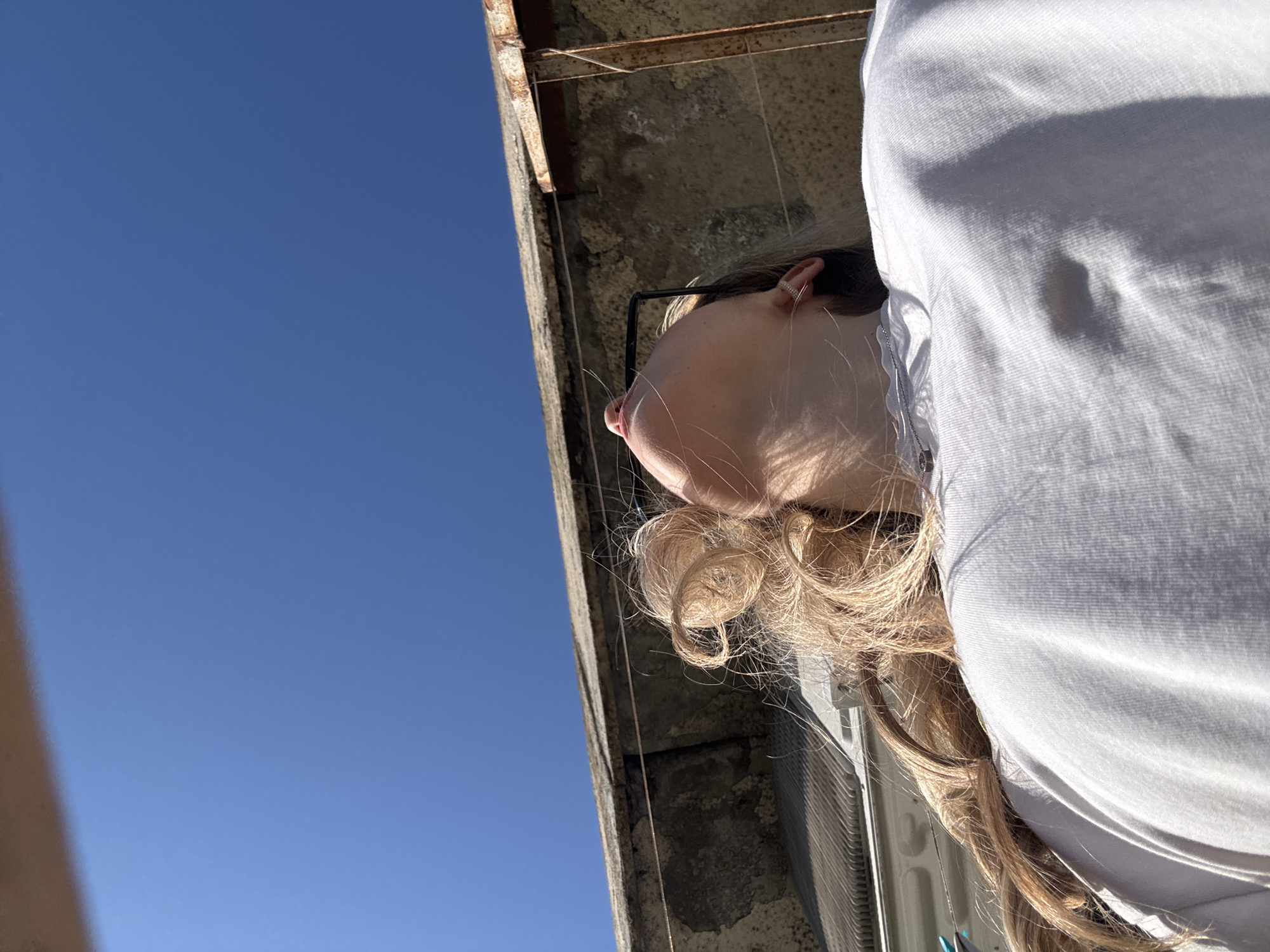 A low-angle, upward shot depicts a woman with blonde hair standing on what appears to be a rooftop or balcony in an urban environment. The background features a clear blue sky. A concrete ledge or part of the building structure is visible near the woman. She is wearing a light-colored shirt. The location is described as Unknown, Unknown.