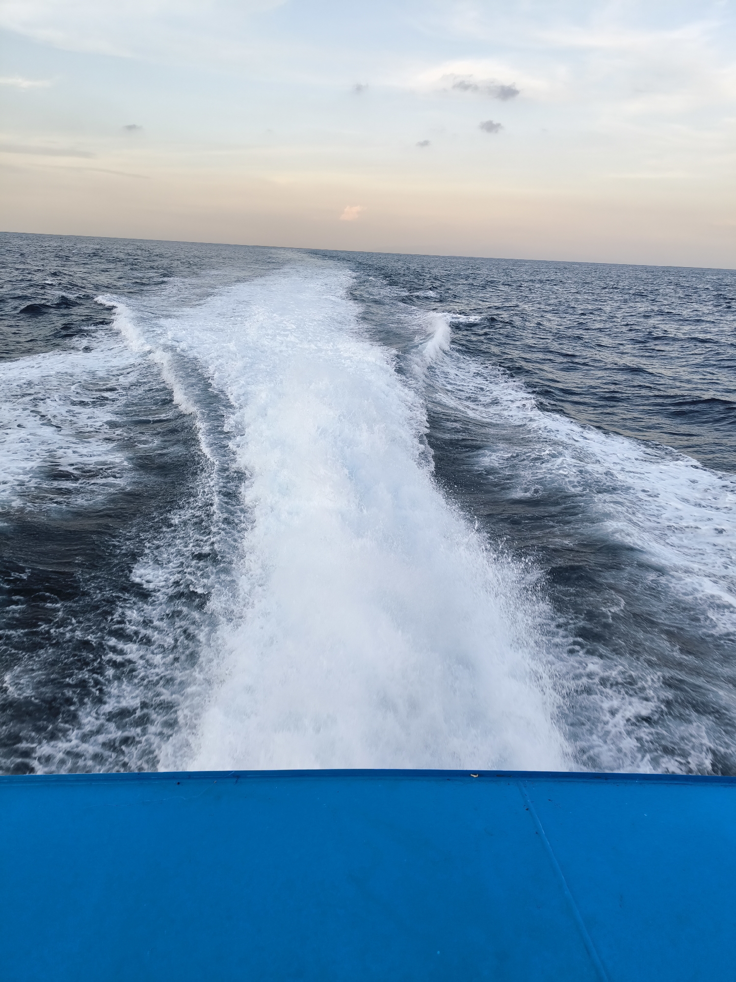 A photograph taken on a boat in Unknown, Indonesia, shows the churning white wake of the vessel moving away from the observer. The blue of the boat's stern is prominent in the foreground. The sea is a darker blue, and the sky is overcast with muted tones. No people are visible.