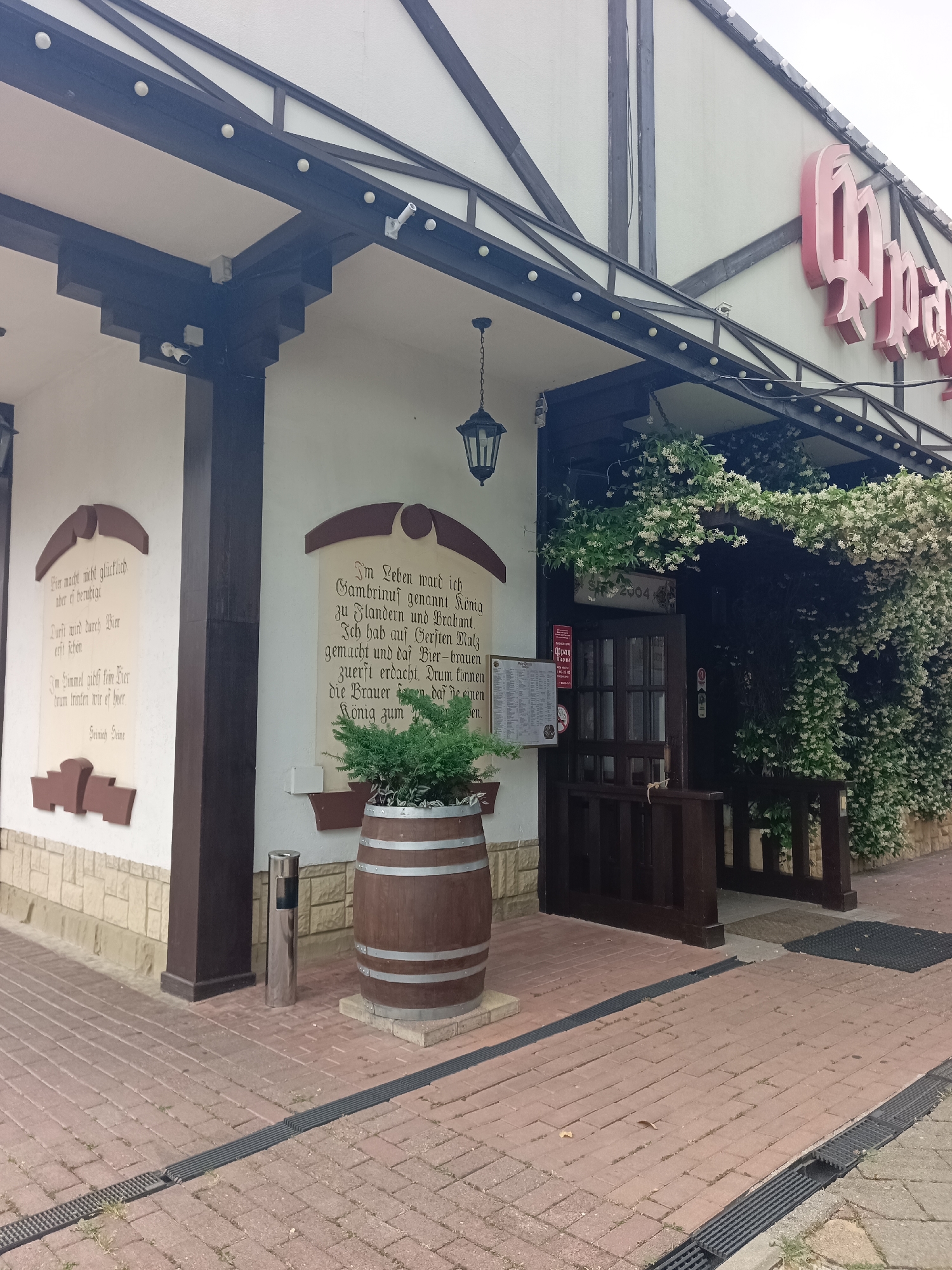 The image depicts the exterior of a restaurant, possibly a German-themed establishment, with a distinctive Tudor-style architectural influence. The building features a white facade with dark brown timber framing, reminiscent of half-timbered construction. Above the entrance, large, bright pink lettering spells out the restaurant's name, which appears to be "Oppa". The entrance itself is a wooden door, partially obscured by abundant white flowering vines that cascade over the awning, creating a lush, verdant archway. A sign above the entrance indicates the year "2004".
To the left of the entrance, a decorative barrel planter holds a small evergreen plant. Two large, framed plaques with text are mounted on the building's wall. The plaque on the left contains German text, and the plaque on the right also features German text and what appears to be a stylized depiction of a beer mug. A black wrought-iron lantern hangs from the overhang.
The ground in front of the restaurant is paved with reddish-brown bricks, arranged in a pattern. A metal trash receptacle stands near the barrel planter. Dark metal grates, likely for drainage, run along the edges of the paved area. The overall impression is one of a charming and inviting establishment, possibly located in a well-maintained area. The presence of the signage and architectural style suggests a specific cultural theme for the restaurant. The image is taken from a slightly low angle, looking upwards towards the building.
