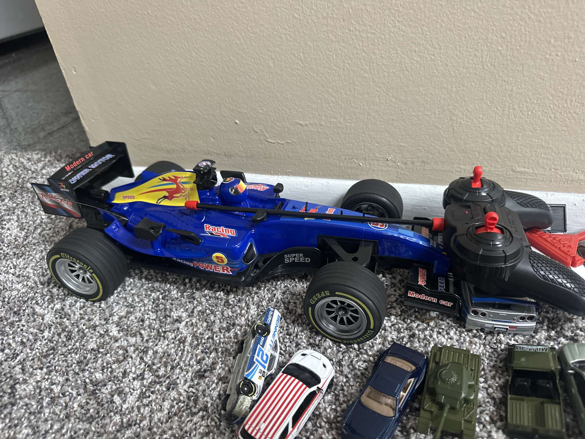 A blue and yellow remote-controlled race car is the primary subject of the image, positioned on a gray carpet. The car features various decals and branding, including "Modern car," "Racing POWER," "SUPER SPEED," and a graphic of a deer. It has black tires with yellow accents and a driver figure in the cockpit. A black remote control with red buttons sits on top of the car. In the foreground, there are several smaller toy vehicles, including a blue and white race car with the number 21, a white car with red stripes, a dark blue car, a green toy tank, a green military truck, and a sliver of a black car. The background consists of a light-colored wall. The scene is indoors and appears to be in a residential setting, possibly a playroom or living area. The image was taken in Schaumburg, United States of America.