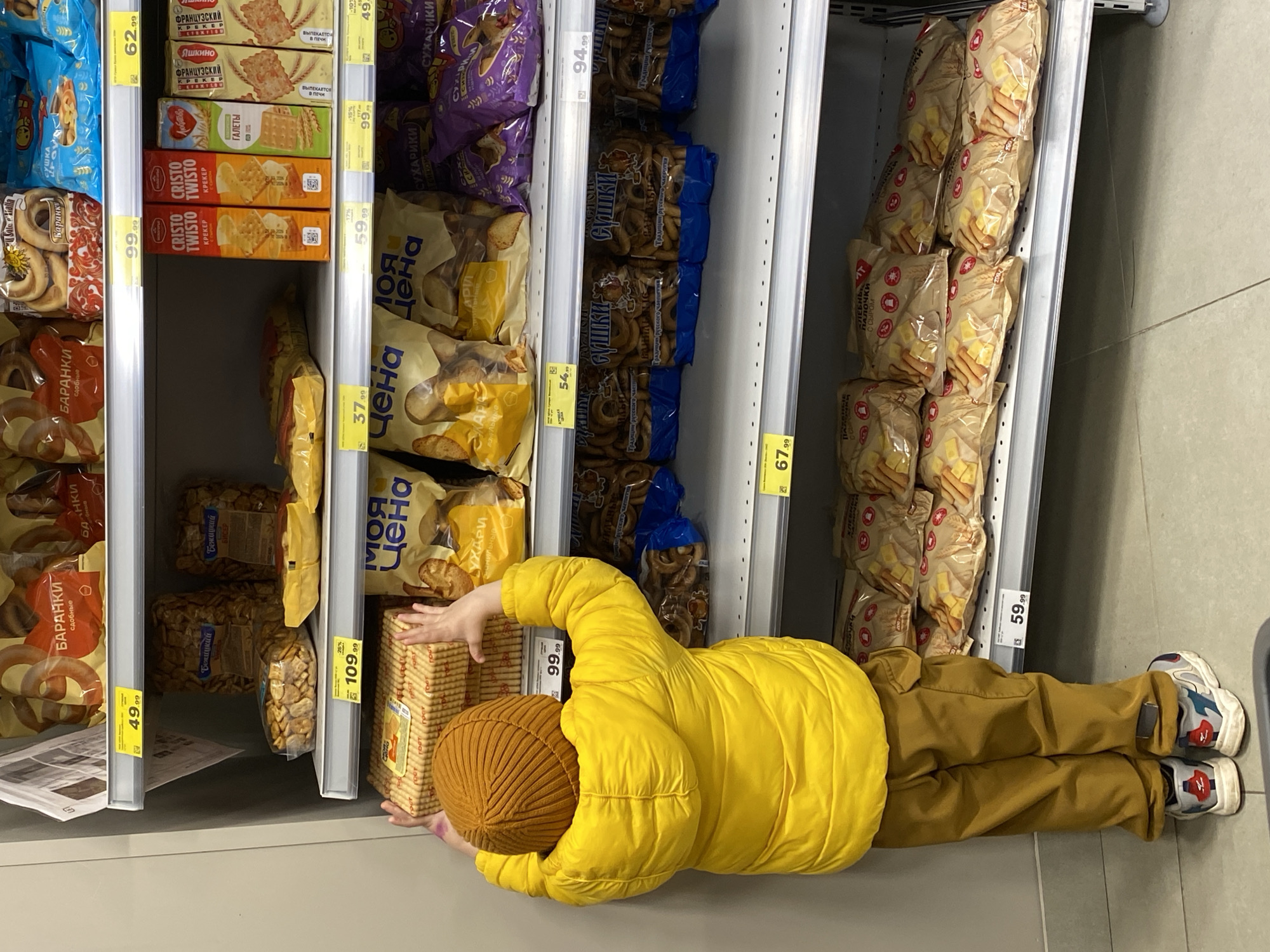 A child in a yellow puffer jacket and brown pants lies prone on the floor of a grocery store in Sochi, Russia, reaching for a box of cookies on a shelf. The child also wears a brown knit hat and sneakers. The shelves are stocked with various packaged baked goods, including crackers, breadsticks, and bagels, with price tags in Russian currency visible. The floor is light gray.
