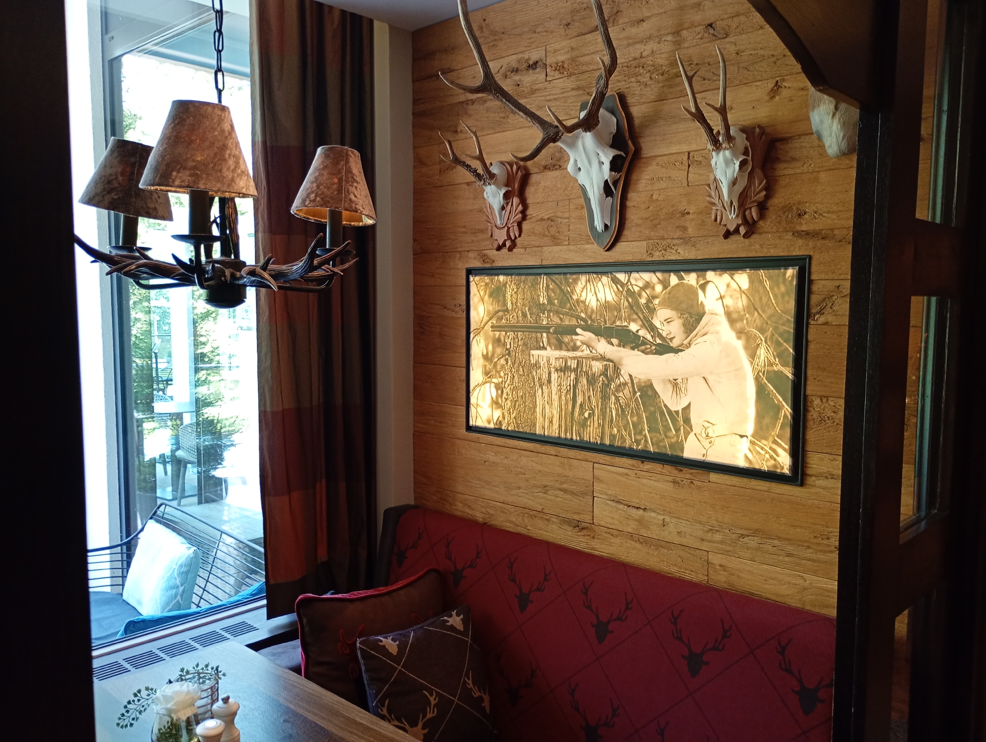 This image depicts an indoor scene, likely a rustic dining area, possibly in a hotel or restaurant in Bad Sachsa, Germany. The decor features a strong hunting or woodland theme.
Dominating the wall is a large, framed picture of a person, likely a young man, aiming a rifle. Above this picture, the wall is adorned with two mounted deer skulls with antlers. The wall itself is paneled with wooden planks.
In the foreground, a deep red upholstered bench or sofa is visible, decorated with a pattern of black deer heads and antlers. Two throw pillows rest on the bench; one is dark brown with a diamond pattern and antler motifs, and the other is a deeper red with a white embroidered motif, possibly a stag's head.
To the left, a large window offers a view of an outdoor seating area with chairs and tables, suggesting a connection to an exterior space. A multi-bulb chandelier with fabric lampshades hangs near the window. The chandelier's base appears to be made of antler-like material.
A wooden table is partially visible in the lower foreground, set with a salt and pepper shaker and a small glass containing greenery and a white flower. The overall ambiance suggests a cozy, traditional, and perhaps hunting-themed establishment.