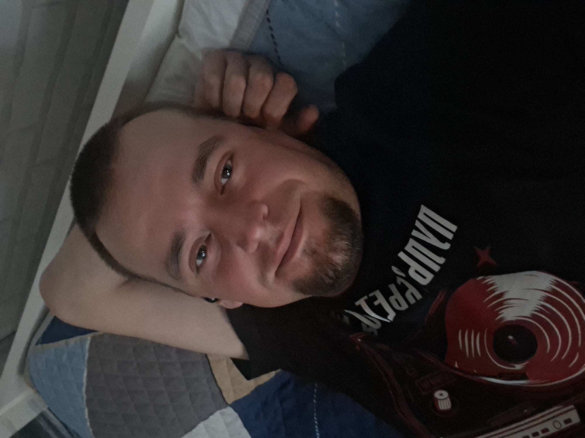 A man is lying down on a bed. He is looking at the camera and smiling. The man has short brown hair and a goatee. He is wearing a black t-shirt. The bed has a blue and gray quilt on it. The room is dimly lit.