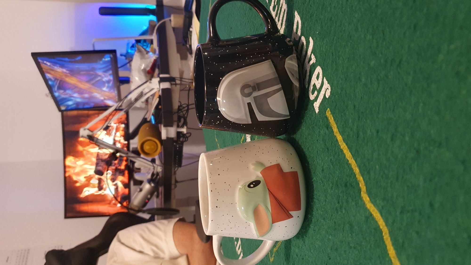 The image shows two mugs, one white with a Grogu (Baby Yoda) design and the other black with a Mandalorian helmet design, sitting on a green surface with "Poker" printed on it. In the background is a desk with two monitors displaying a game and a fire. Also visible is a microphone setup. The location cannot be determined from this image. The time of day cannot be determined.