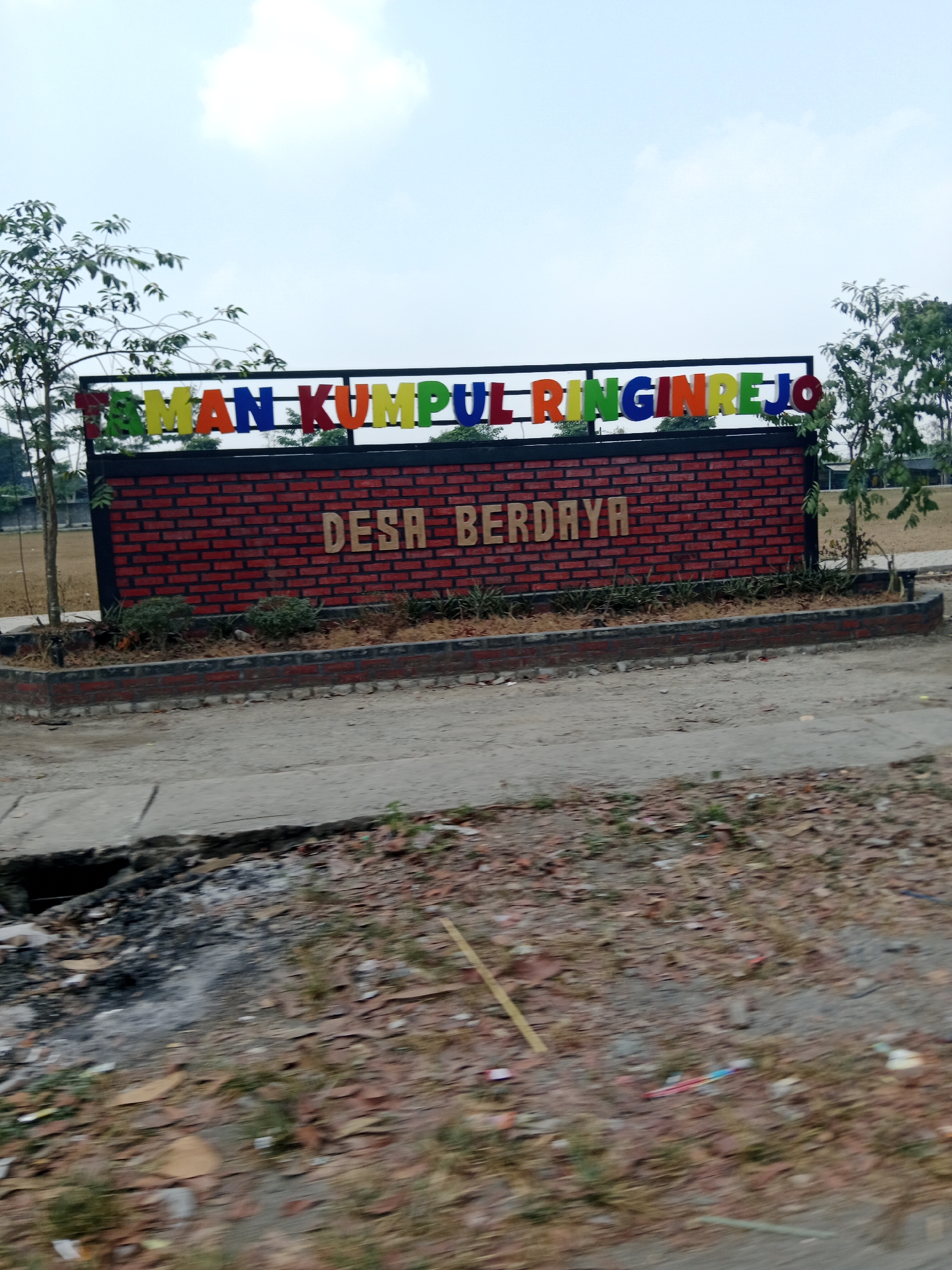 The image shows a sign in front of a brick wall. The sign has colorful letters that spell out "TAMAN KUMPUL RINGINREJO". Below this, a second line of text in gold letters reads "DESA BERDAYA". The brick wall is decorated with green plants at its base. In the foreground, there is a dirt path with some leaves and debris. The background shows trees and a cloudy sky. The overall impression is of a welcoming entrance to a park or public space in the village of Ringinrejo, Indonesia.