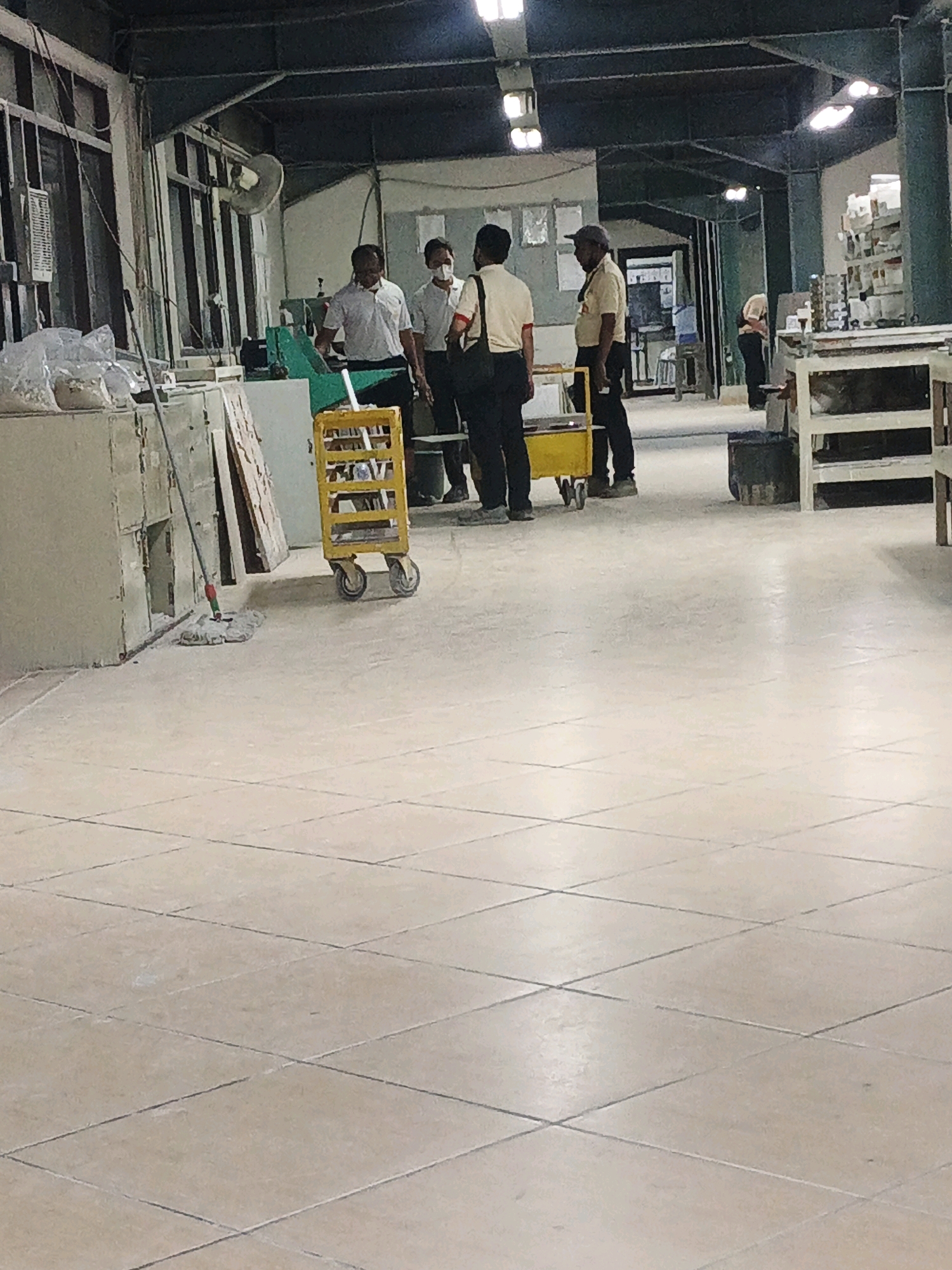 The image depicts an indoor industrial or workshop environment. In the foreground, a light-colored tiled floor stretches out, showing some wear and tear. Several individuals are visible in the mid-ground, appearing to be engaged in a discussion or work-related activity. One person, wearing a light-colored shirt and dark pants, stands facing away from the camera. To their left, another person in a white shirt and mask, and to the right, a person in a light-colored shirt and dark pants. In the background, there are various pieces of equipment and structures, suggesting a functional workspace. A yellow cart with wheels is positioned in the mid-ground. The overall lighting appears to be artificial, coming from overhead fixtures. The presence of a mop leaning against a wall in the left mid-ground indicates ongoing cleaning or maintenance. The location is identified as Ngoro, Indonesia, implying this is a scene from a business or factory within that area.