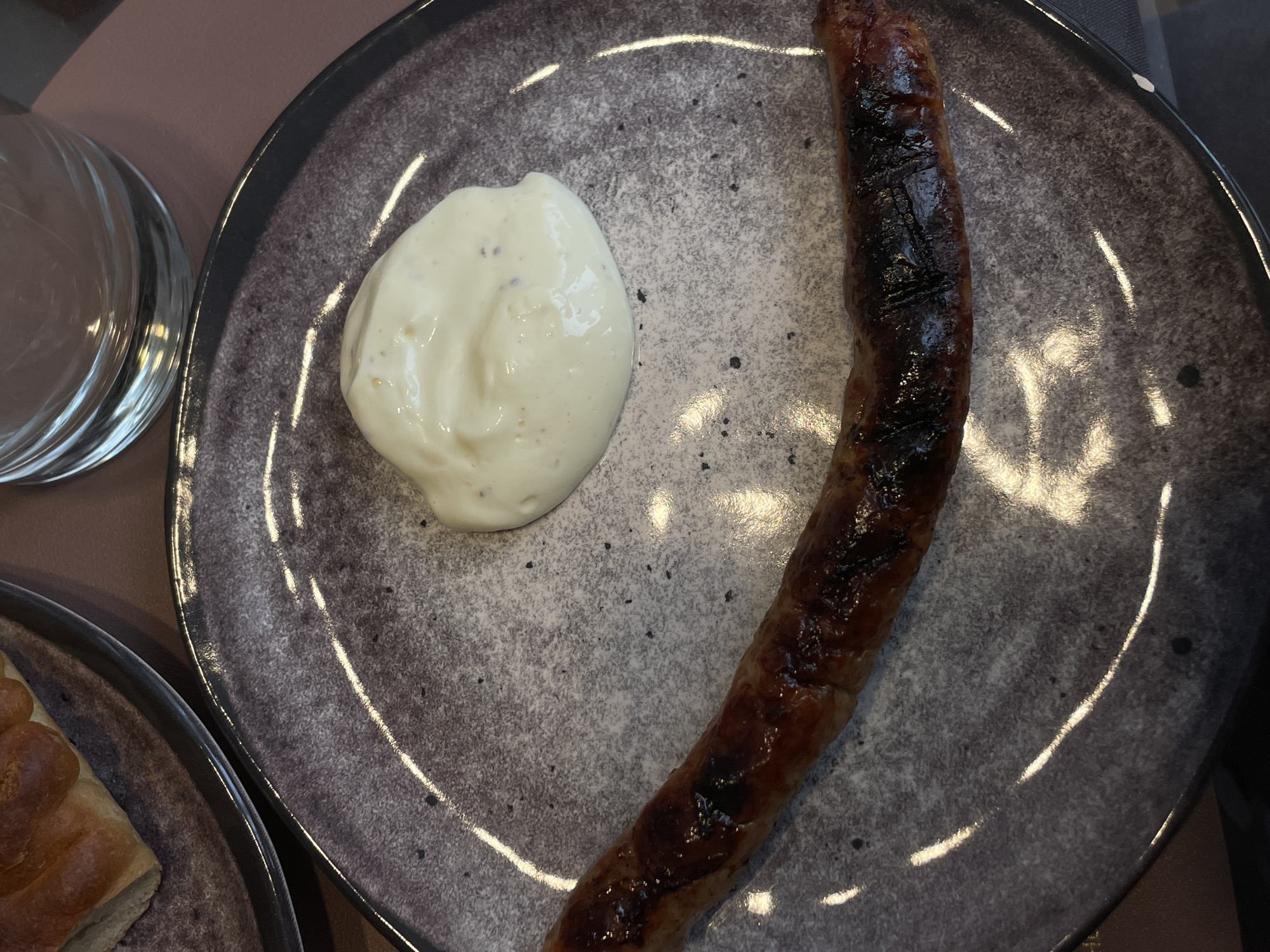 A plate contains a grilled sausage and a dollop of white sauce. A glass and another plate with food are partially visible. The image does not provide information to determine if the location is Подшивалово, Russia.