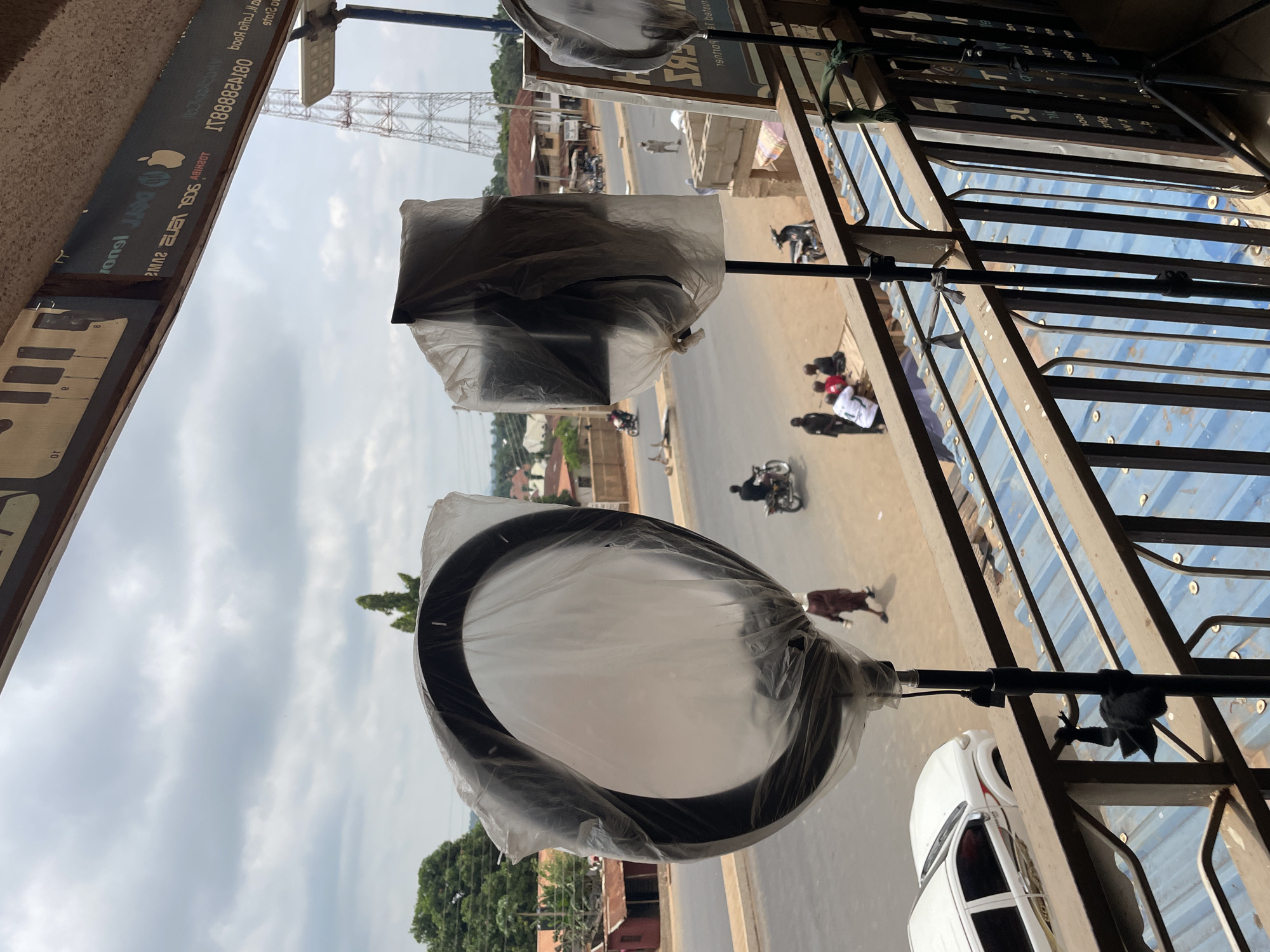 This image, likely taken from a shop in Akwanga, Nigeria, shows a street scene viewed through an opening in a building. Visible in the foreground are two light fixtures still wrapped in protective plastic, hanging over the edge of a balcony or window. Below, a road runs through what appears to be a bustling area, with pedestrians, motorcycles, and at least one white car. A communications tower is visible in the distance, indicating the presence of telecommunications infrastructure. The sky is overcast with cloud cover.
