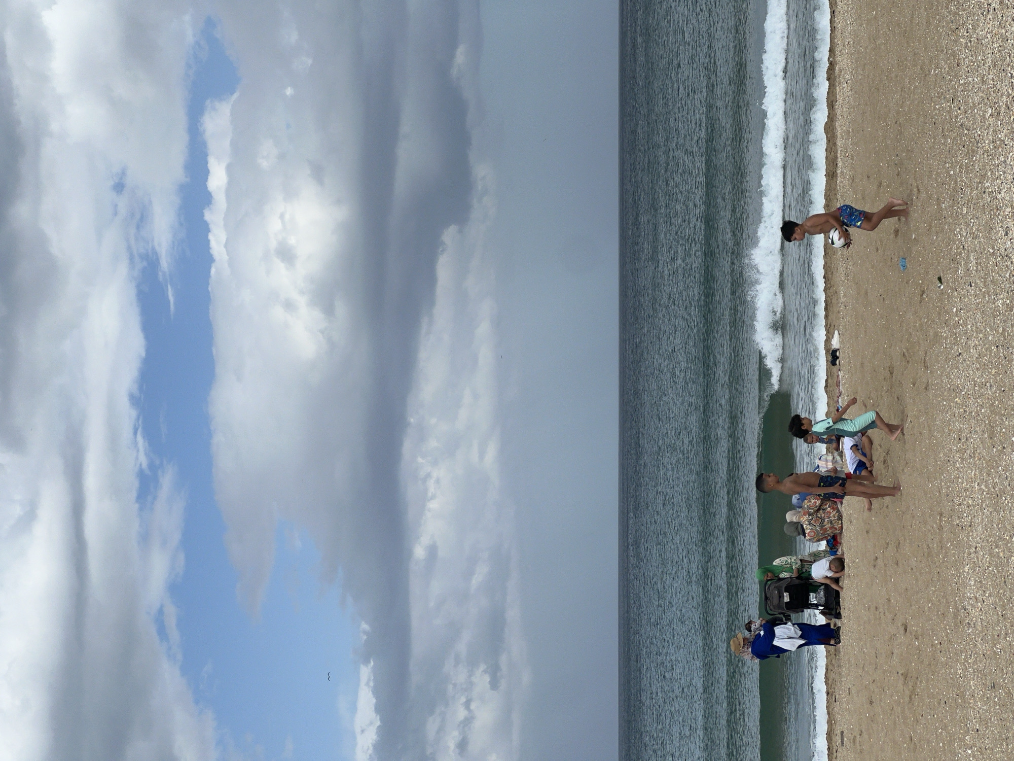 The image shows a beach scene in M'Diq, Morocco. The sky is partly cloudy. The sea is adjacent to the beach, with waves visible. A group of people is on the beach. One boy is running. Another boy with a ball. Others are standing, sitting, or pushing a stroller. The beach is sandy with some shell fragments.