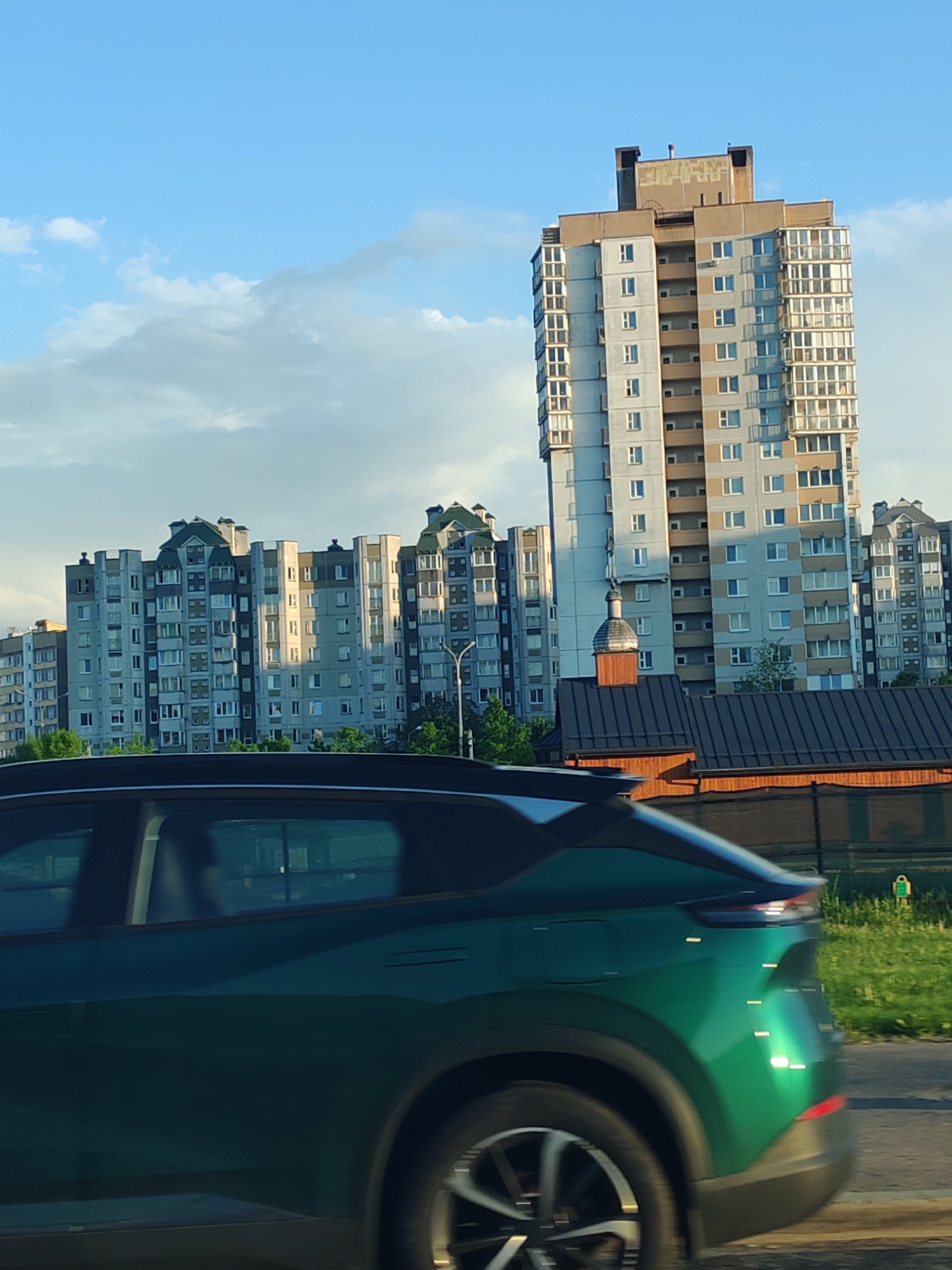 A view from within a moving car shows a series of multi-story buildings under a partly cloudy sky. A tall, rectangular building stands out with its gray and brown facade. A church with a small onion dome is visible behind a dark-roofed structure. In the foreground, a green vehicle is in motion, obscuring the lower part of the view. Patches of grass and a fence line the edge of the road. The image is taken in the city of Unknown, Unknown.