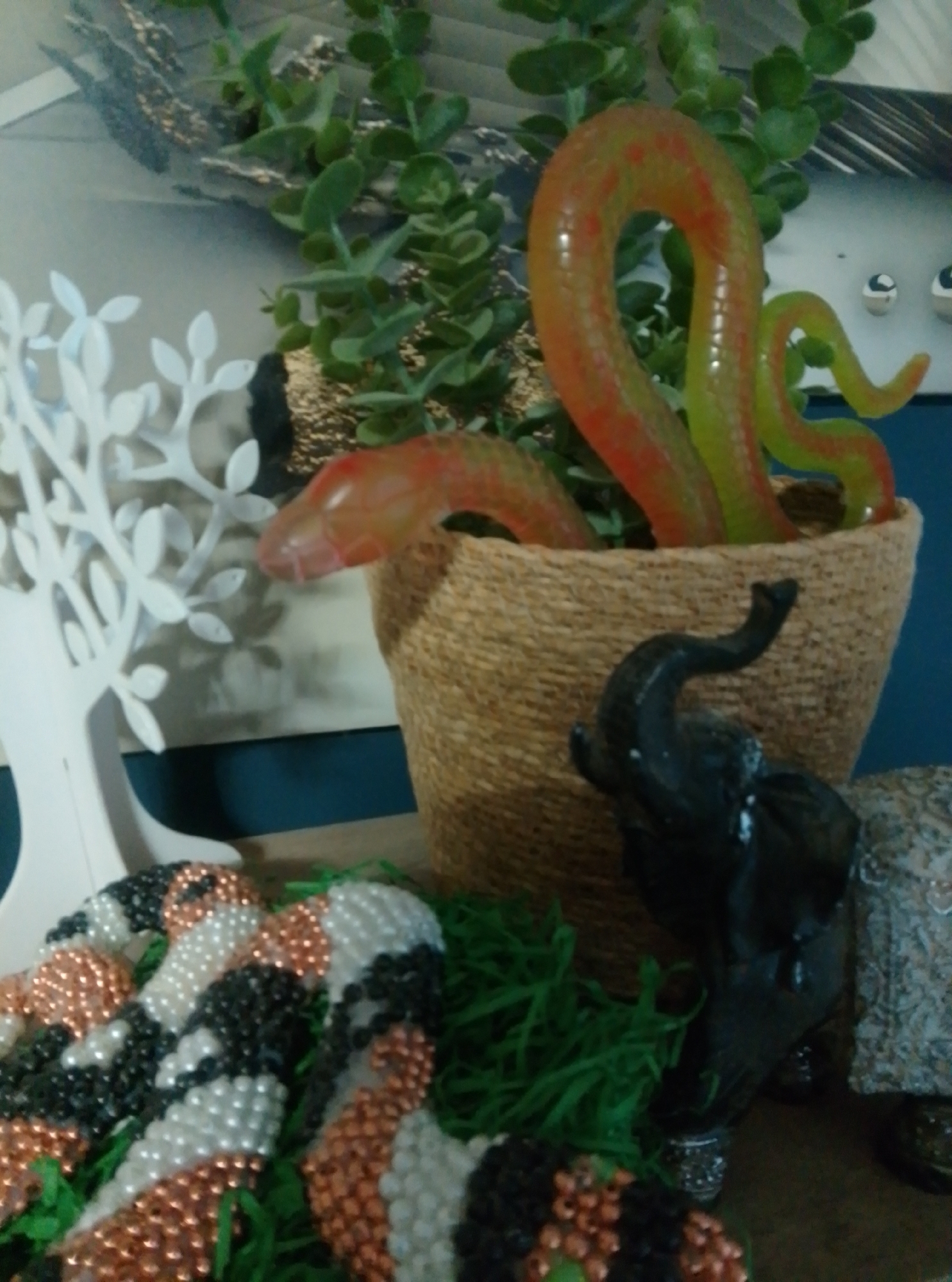 The image depicts a still life arrangement of various decorative objects. In the foreground, a beaded snake-like object, rendered in black, white, and copper beads, lies coiled on green artificial grass. To the right of this beaded snake is a black figurine of an elephant, its trunk raised. Behind these, a woven basket holds a translucent, segmented snake-like sculpture in shades of orange and green. To the left, a white, stylized tree sculpture with leaf-like branches stands against a blurred background. The overall setting appears to be indoors, possibly a display or a shelf arrangement. The location in Wedel, Germany, is not discernible from the visual information presented in the image.
