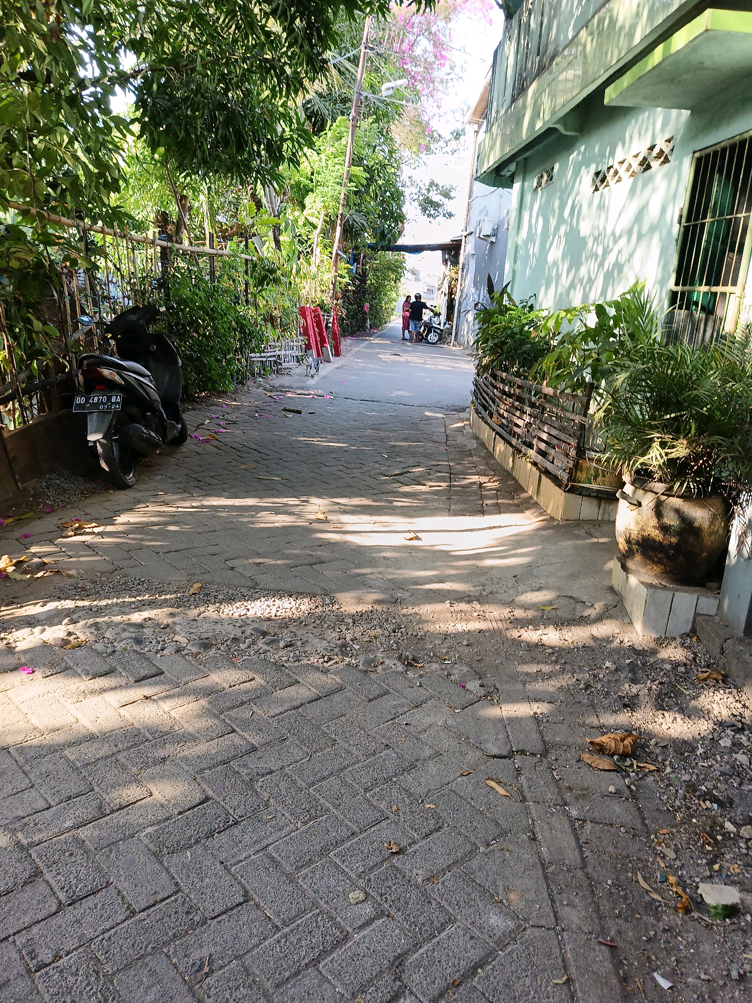 The image depicts a narrow, paved alleyway in Makassar, Indonesia. The alley is lined with lush green foliage on the left, with a dark-colored scooter parked near the beginning of the path. The paving stones are laid in a herringbone pattern, with some areas showing signs of wear and tear. On the right side of the alley, a light green building with barred windows and ventilation blocks is visible. A wooden fence separates the building from the alley, and a large, dark pot with a green plant sits on a concrete base. The scene is illuminated by dappled sunlight, casting shadows across the path. Two individuals, a man and a woman, are seen walking away from the camera further down the alley, near a second scooter. The overall impression is of a residential or commercial side street, typical of many urban areas in Indonesia.