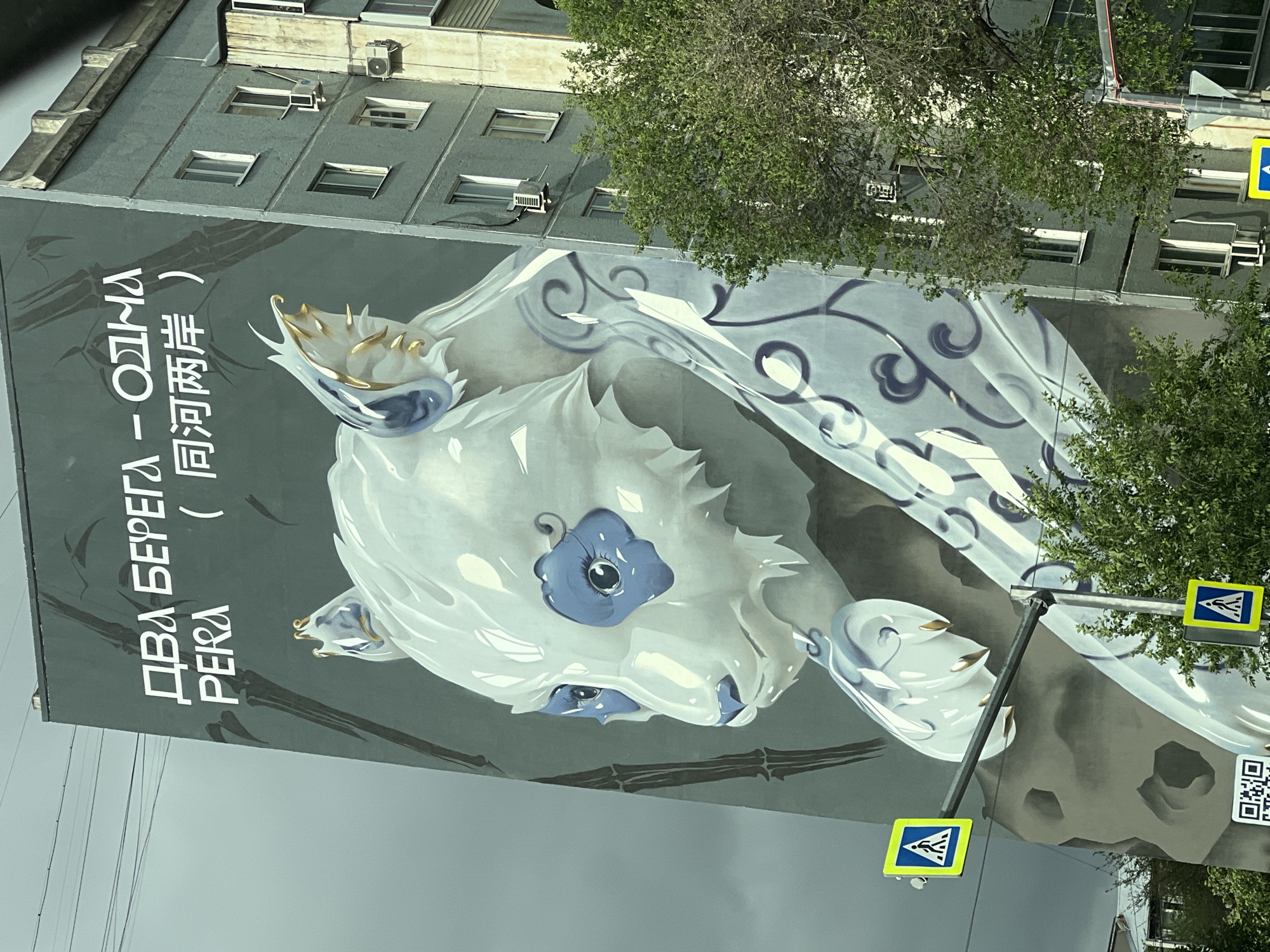 A partial, canted, aerial view depicts the side of a gray building with a large mural. The mural features an image of a white, stylized animal head with blue accents, possibly a panda or similar creature. The head has swirling designs around it, giving the impression of movement or energy. Below the animal, there are horizontal gray bamboo stalks. To the left of the mural, there are vertical lines of white text written in both Cyrillic and Chinese characters.
The building itself has several windows and air conditioning units. Parts of green trees obscure the mural and the building. A pole with two blue pedestrian crossing signs is visible in front of the mural, and a QR code is present on the right side. The sky is overcast. The location is presumed to be the city of Unknown, Unknown based on the prompt.