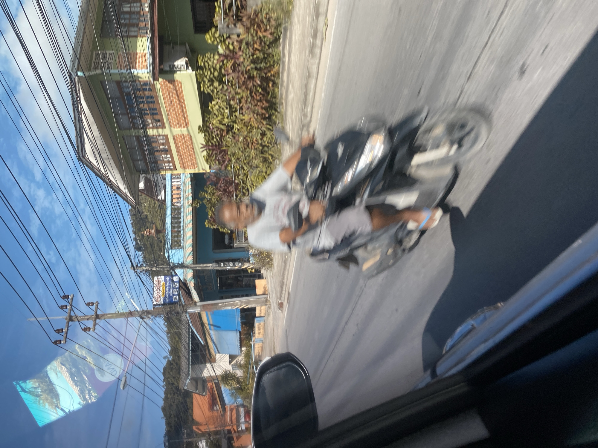 A man is riding a scooter on a street in Baan Hua Thanon, Thailand. He is wearing a light-colored t-shirt and shorts, and white flip-flops. The scooter is black. The man is in motion, and the background is blurred. The background consists of buildings, trees, and a blue sky with some clouds. There are power lines visible across the sky. The image appears to have been taken from inside a vehicle.