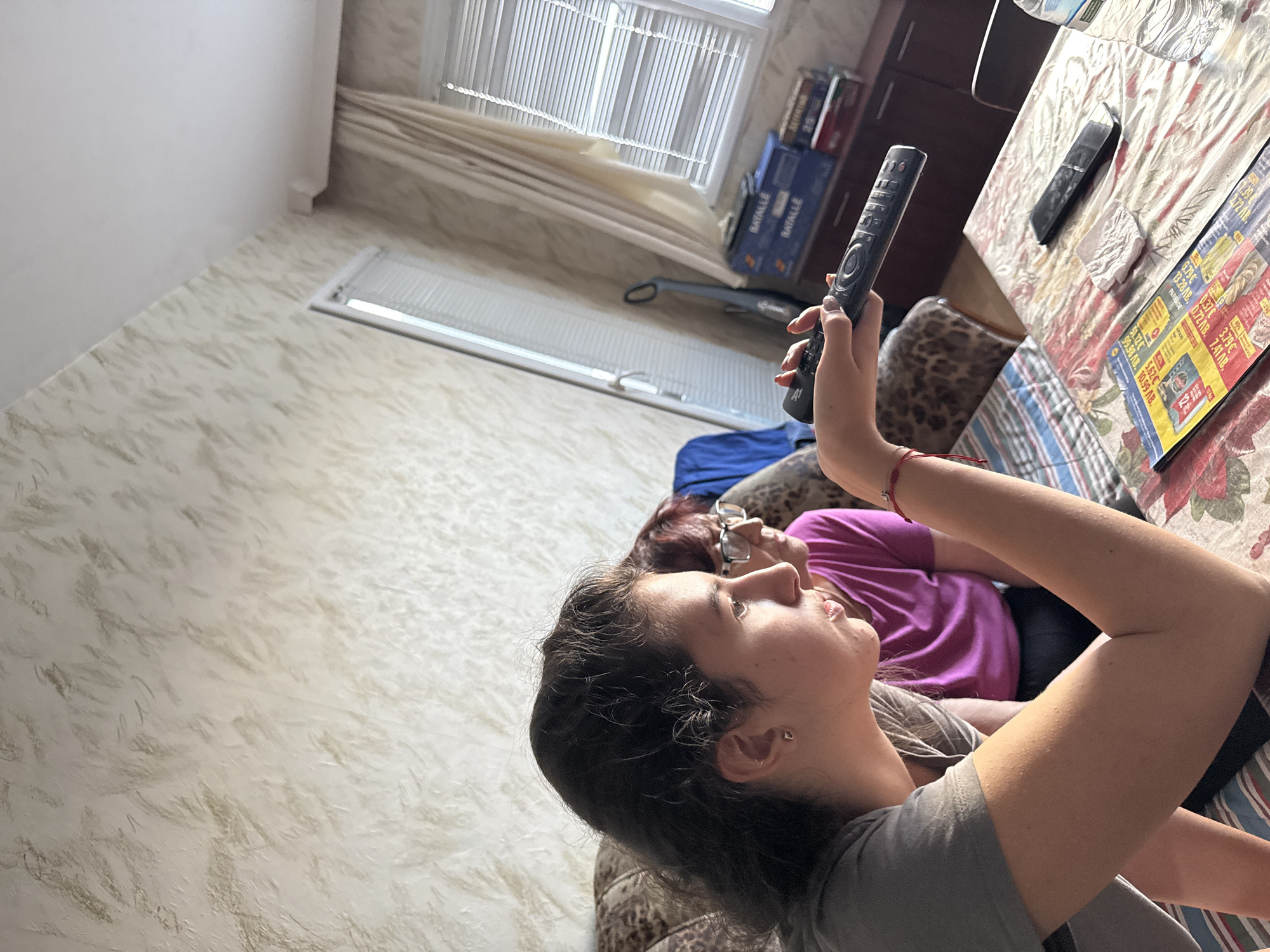 The image depicts an indoor residential scene in what appears to be Stara Zagora, Bulgaria, featuring two women. In the foreground, a young woman with dark, wavy hair and a grey t-shirt is presented in profile, looking towards the right of the frame. Her right hand, adorned with light-colored nail polish, firmly grasps a black remote control, pointed in the same direction. A red string bracelet is visible on her left wrist. Behind and to her left, an older woman with reddish hair and glasses is partially visible, wearing a purple t-shirt, also oriented towards the right. Both women are seated on a couch featuring a brown and black patterned upholstery.
A table in front of the young woman is covered with a floral-patterned tablecloth. On this table, a folded advertisement or flyer is prominently displayed, showing text and prices in Bulgarian Lev (лв), such as "0.99 лв." and "2.79 лв.". Additionally, a second black remote control, a crumpled white napkin, and a partially visible plastic water bottle are present on the table.
The background showcases a room with light beige, textured walls. A large white-framed window or balcony door, fitted with horizontal blinds, is visible, accompanied by a light-colored curtain drawn to its right. A grey stick vacuum cleaner stands upright near the window. Further to the right, a dark brown cabinet or shelf unit contains multiple blue boxes labeled "BATALLIE" and a stack of books or other items. The ceiling is white. The scene is illuminated by natural light, indicating daytime.