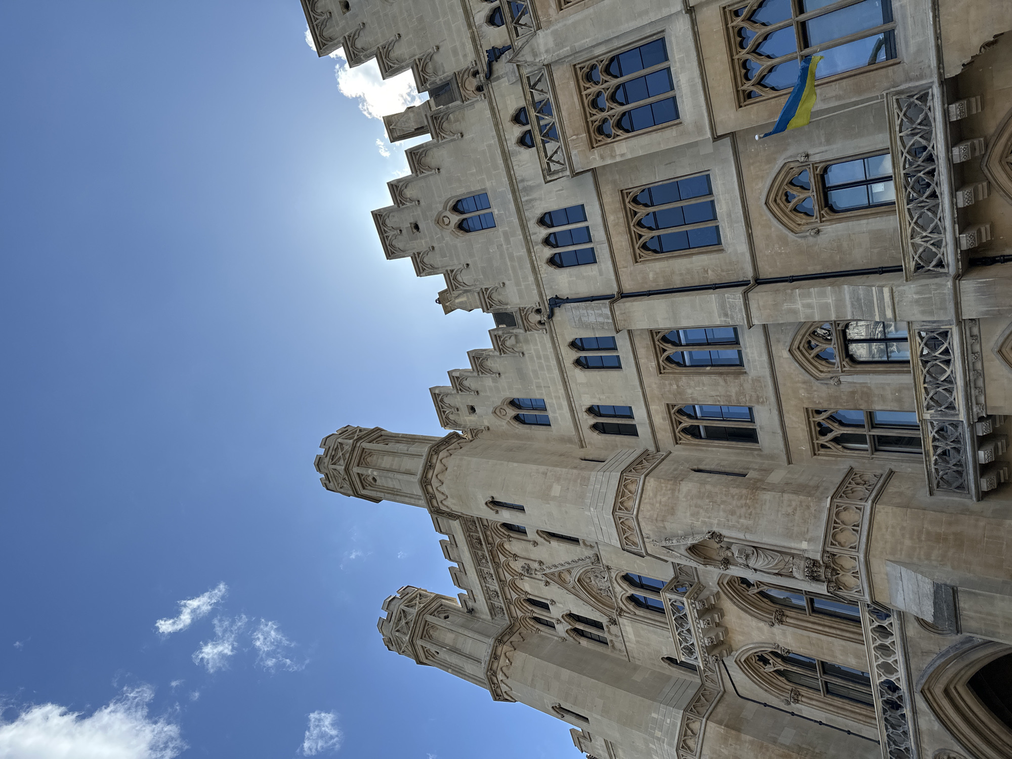 The image showcases a building in the city of London, United Kingdom, oriented from a low-angle perspective. The architectural style is reminiscent of Gothic Revival, featuring elaborate stonework, pointed arches, and decorative battlements. The facade is predominantly light beige in color and includes rows of windows with detailed tracery. A flag featuring blue and yellow horizontal bands, resembling the Ukrainian flag, is visible attached to the building. The sky is blue with scattered white clouds, and the sun is partially obscured by the building, causing a bright glare. No people are visible in the image.