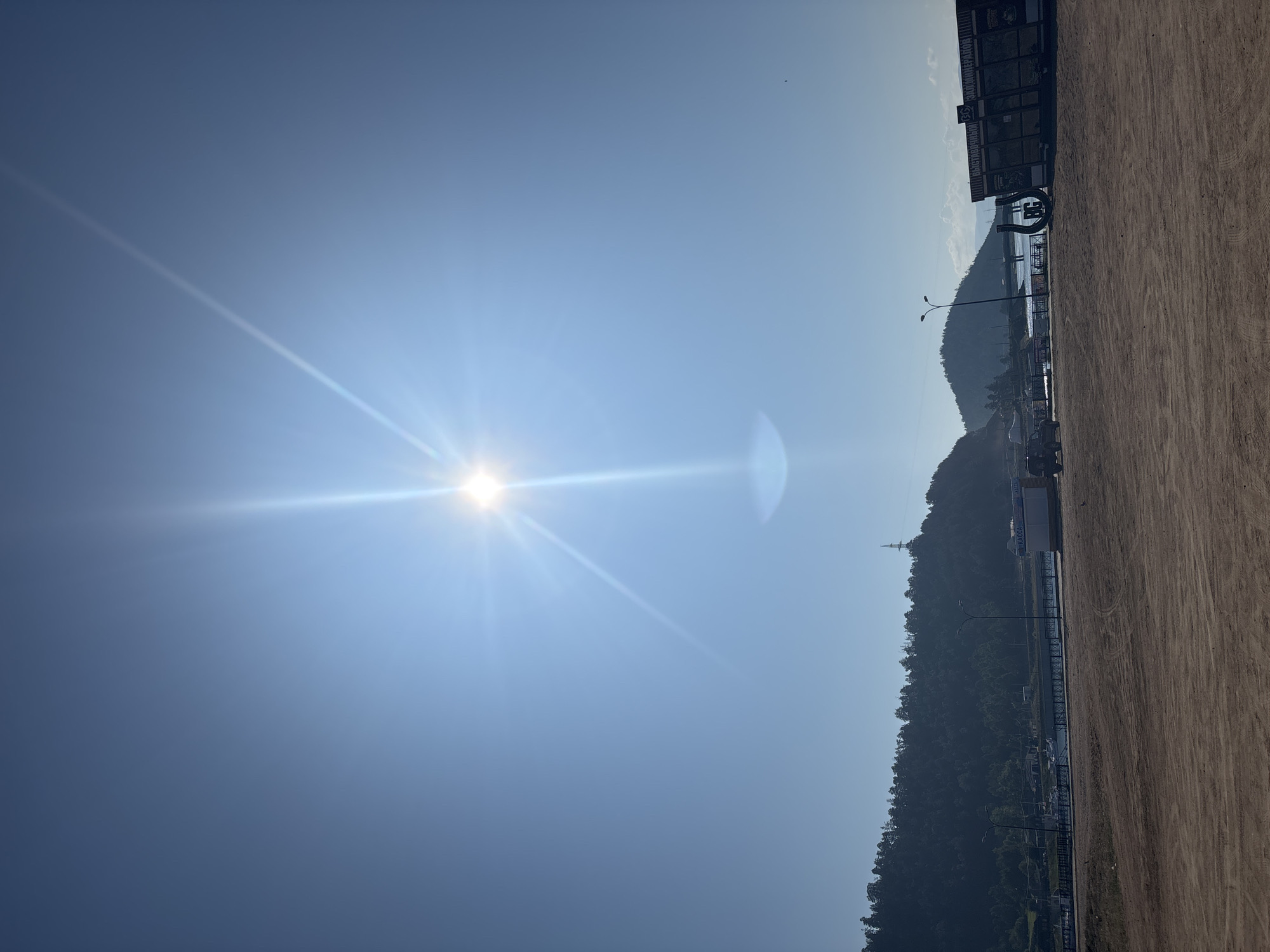 The image depicts a wide, sun-drenched landscape in Turka, Russia, with a clear blue sky dominating the upper portion. A bright sun with prominent rays and a lens flare is positioned in the upper center of the frame. Below the sky, a line of dark green, densely forested hills stretches across the horizon, with a single transmission tower visible on one of the peaks. In the foreground, a vast expanse of dry, sandy ground shows tire tracks, suggesting it is a road or a parking area. Along the shoreline, a body of water is visible, with a few boats moored and a walkway or pier extending into the water. Several buildings, including a small kiosk labeled "КАССА" (KASSA - meaning "cash desk" or "ticket office" in Russian) and a larger structure with signs in Russian, are present along the waterfront. A dark-colored SUV is parked on the sandy ground to the left of the kiosk, and a small tractor-like vehicle is visible near the water's edge. The overall scene conveys a sense of a sunny day in a waterfront area with some commercial activity.
