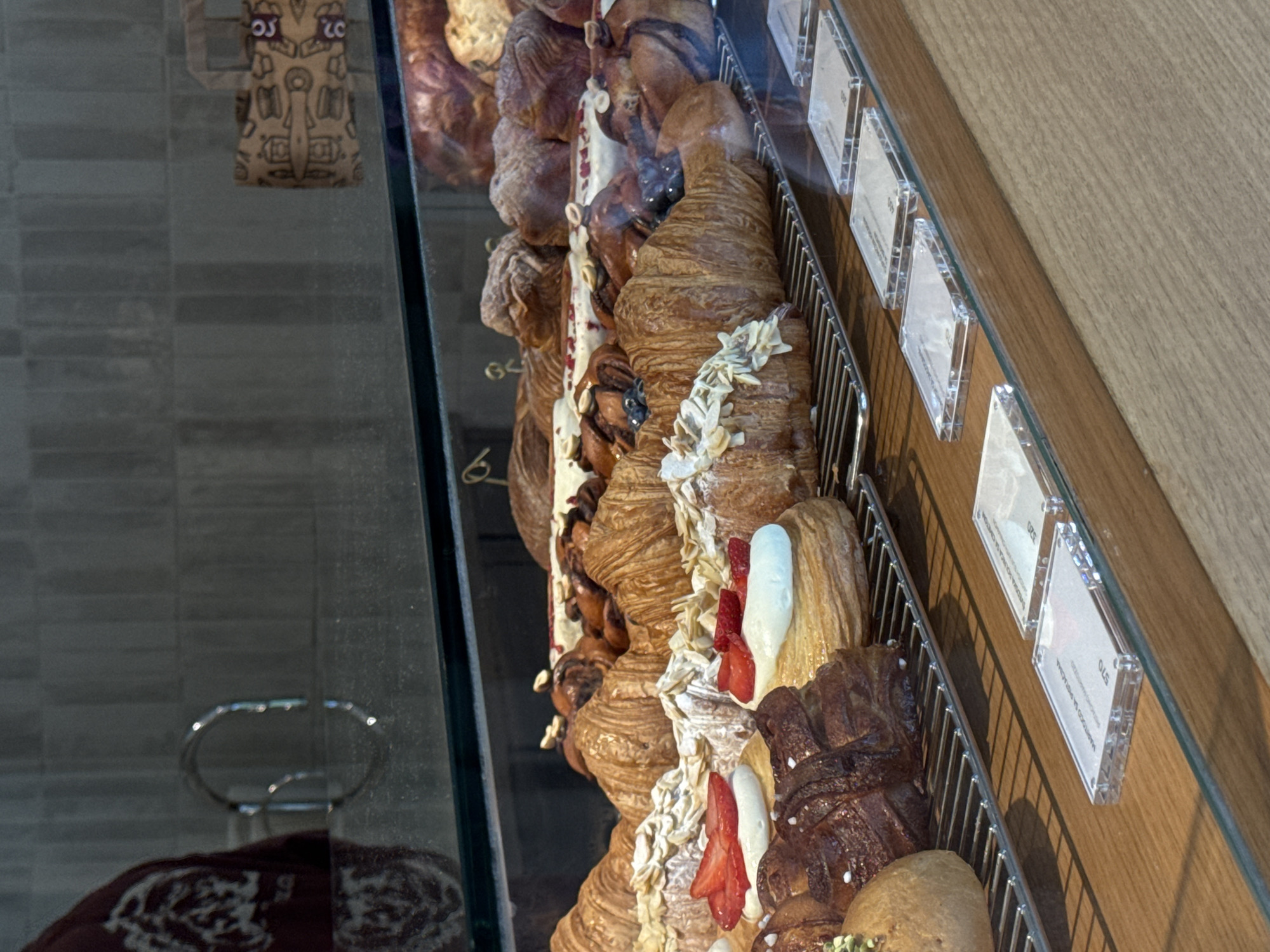 The image depicts an indoor display case filled with various pastries, likely located in Belgrade, Serbia. Croissants and other baked goods are visible, some adorned with toppings such as almonds, cream, and strawberries. Clear price tags are attached to the side of the display, indicating the cost of each item. The display case is made of glass and metal, with a wooden surface adjacent to it. A reflection of interior elements, including a chair and a tiled floor, is visible on the glass surface of the case.