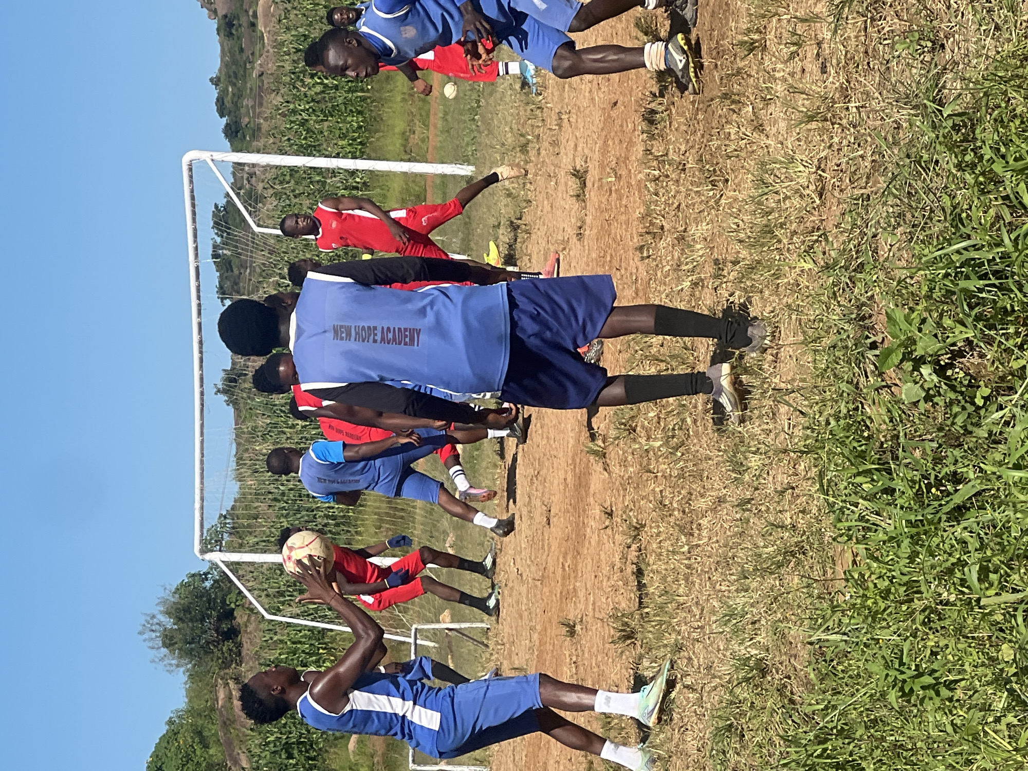 The image shows a group of young men playing soccer on a dusty, outdoor field under a clear blue sky. There is a white goal frame in the background, and the field is bordered by tall green corn stalks and sparse vegetation. Most of the players are wearing athletic shorts and jerseys, with some in blue and white and others in red and white. One player in a blue and white uniform is holding the soccer ball in his hands, appearing to be in the process of throwing it. Another player in a blue uniform with "NEW HOPE ACADEMY" visible on the back is facing away from the camera. The ground is a mix of dry earth and patchy grass. The overall setting suggests a training session or a casual game. This image does not provide any specific visual cues to determine its exact location within Jos, Nigeria.